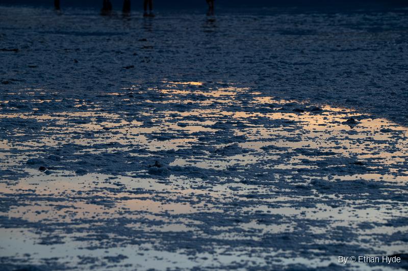 Shallow water pools between clumps of salt crust, reflecting an orange and pale blue sky, with the silhouetted legs of a few people at the top edge.