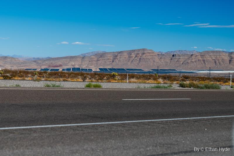 Rows of dark solar panels stretch across desert flats below layered tan ridges, seen past yucca scrub and a paved highway.