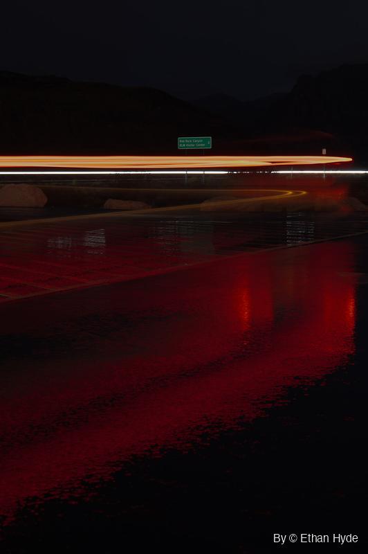 An orange taillight streak and a thin white light trail cross the frame at night above a rain-wet parking area glowing red, with dark mountains behind.