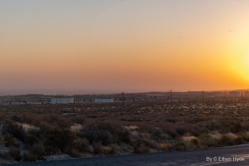 The sun breaks the horizon in an orange sky over desert scrub, with the oil field's tanks and power poles strung along the distance.