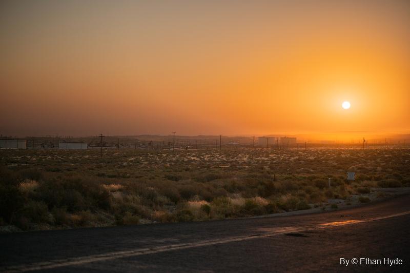 An orange sun rises in haze over desert scrub and the distant McKittrick oil field, a road curving through the foreground.