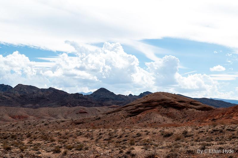 Layered red and tan desert hills in front of dark rocky peaks, under white cumulus clouds and a high sheet of cloud.