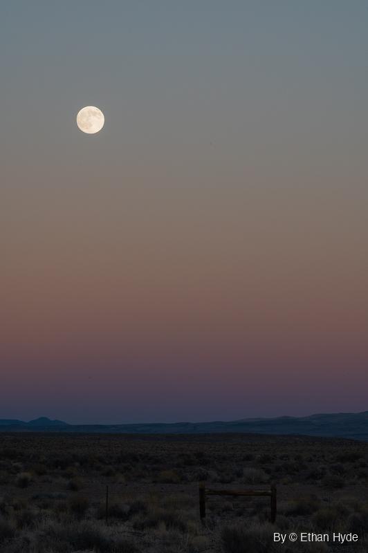 Vertical frame of a near-full moon in a dusk sky fading from blue-gray to pink over distant hills and dark sagebrush, with a wooden fence brace in front.