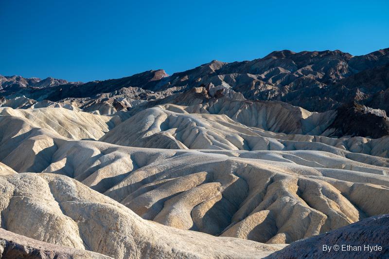 Pale, water-carved badlands of Zabriskie Point roll toward dark mountains under a deep blue sky.