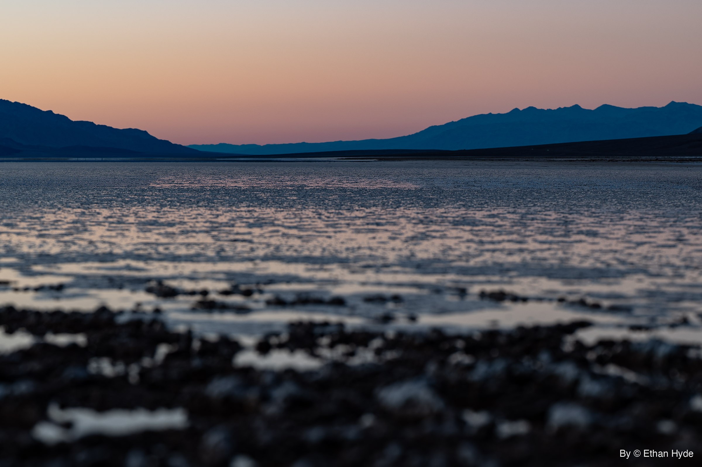 A ground-level view across blurred salt crust and shallow water toward silhouetted mountains under a pink dusk sky.