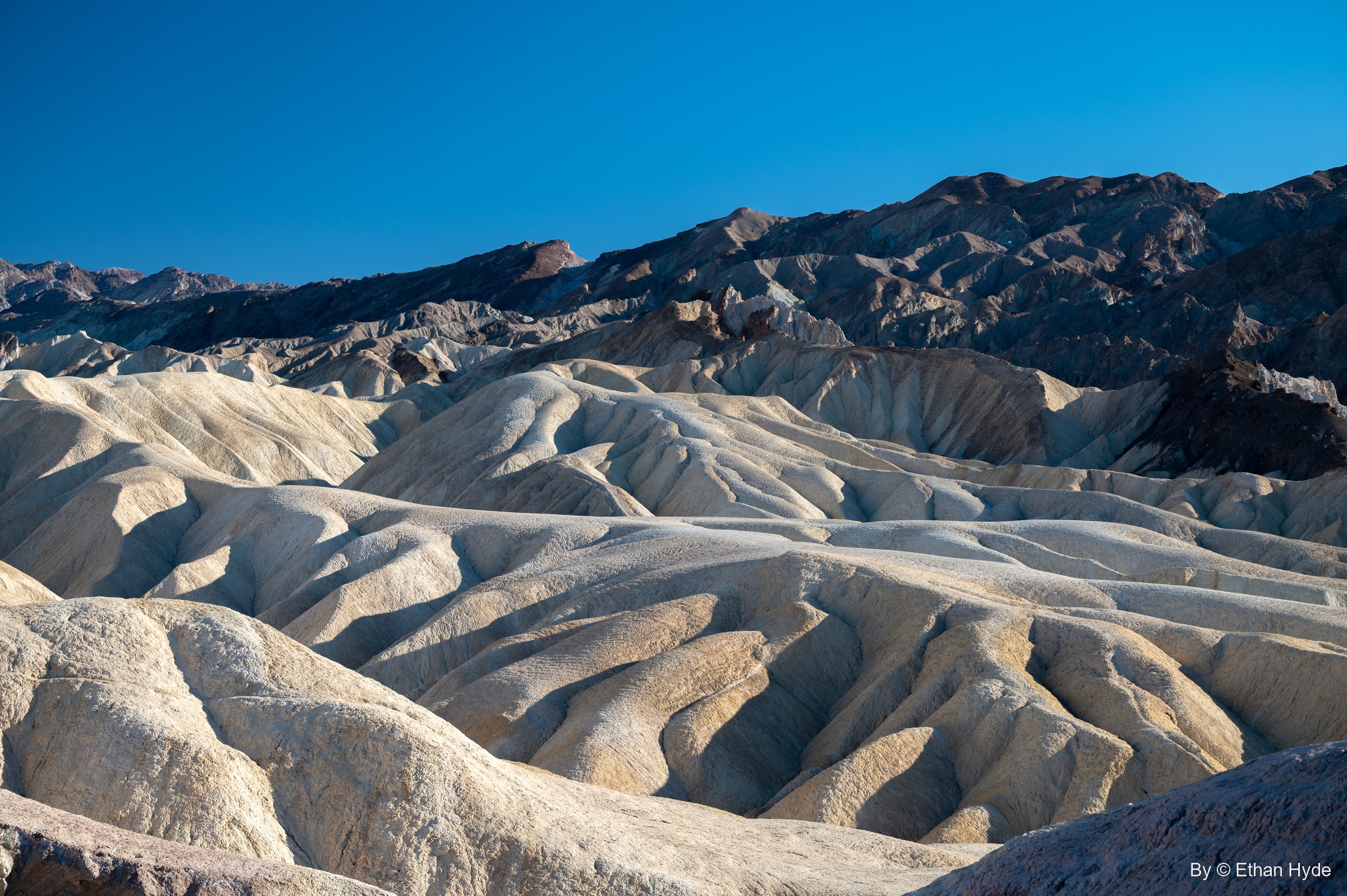 Pale, water-carved badlands of Zabriskie Point roll toward dark mountains under a deep blue sky.