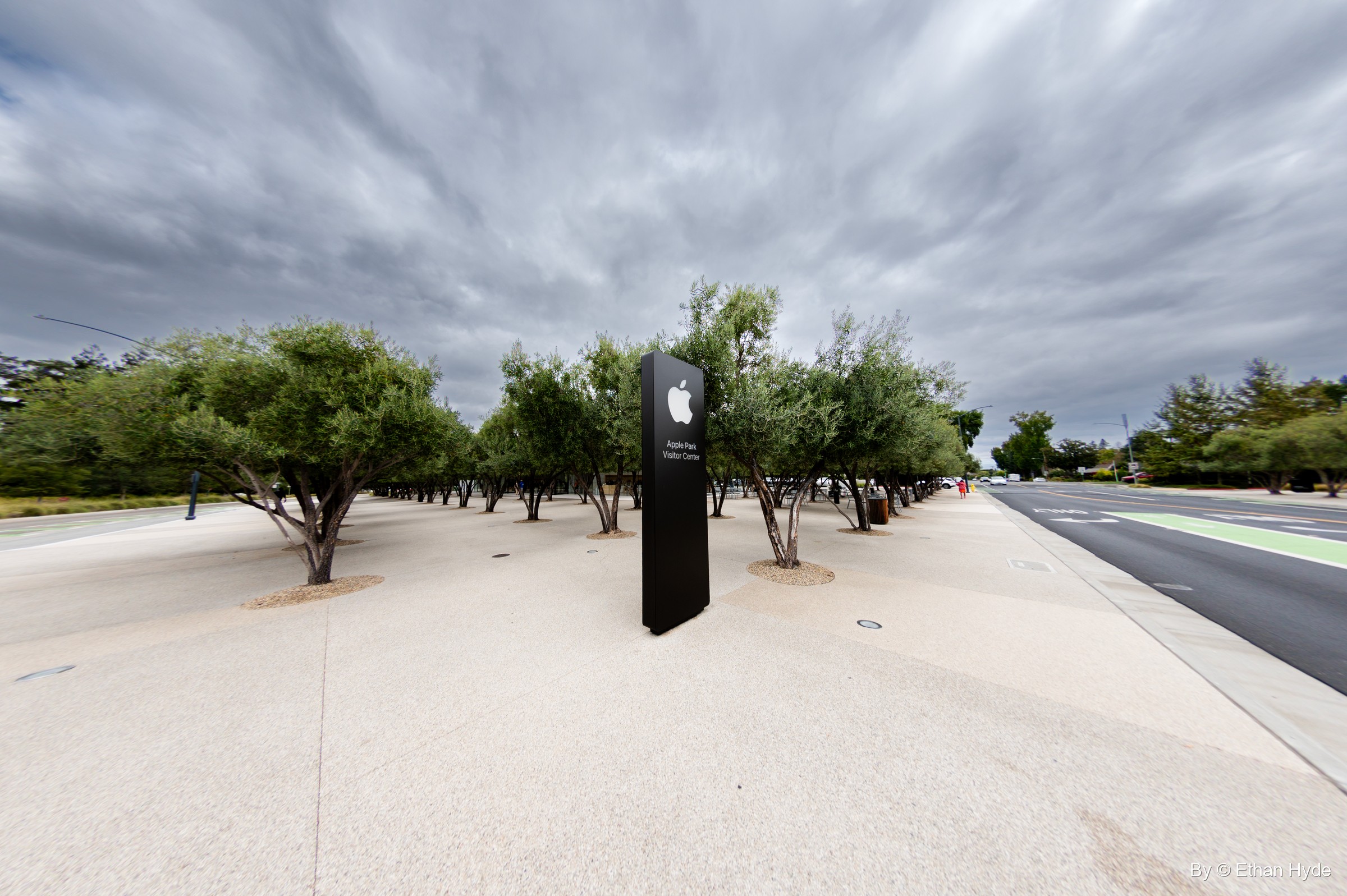 A black Apple logo sign stands among rows of olive trees on a pale plaza beside the road, under gray clouds.