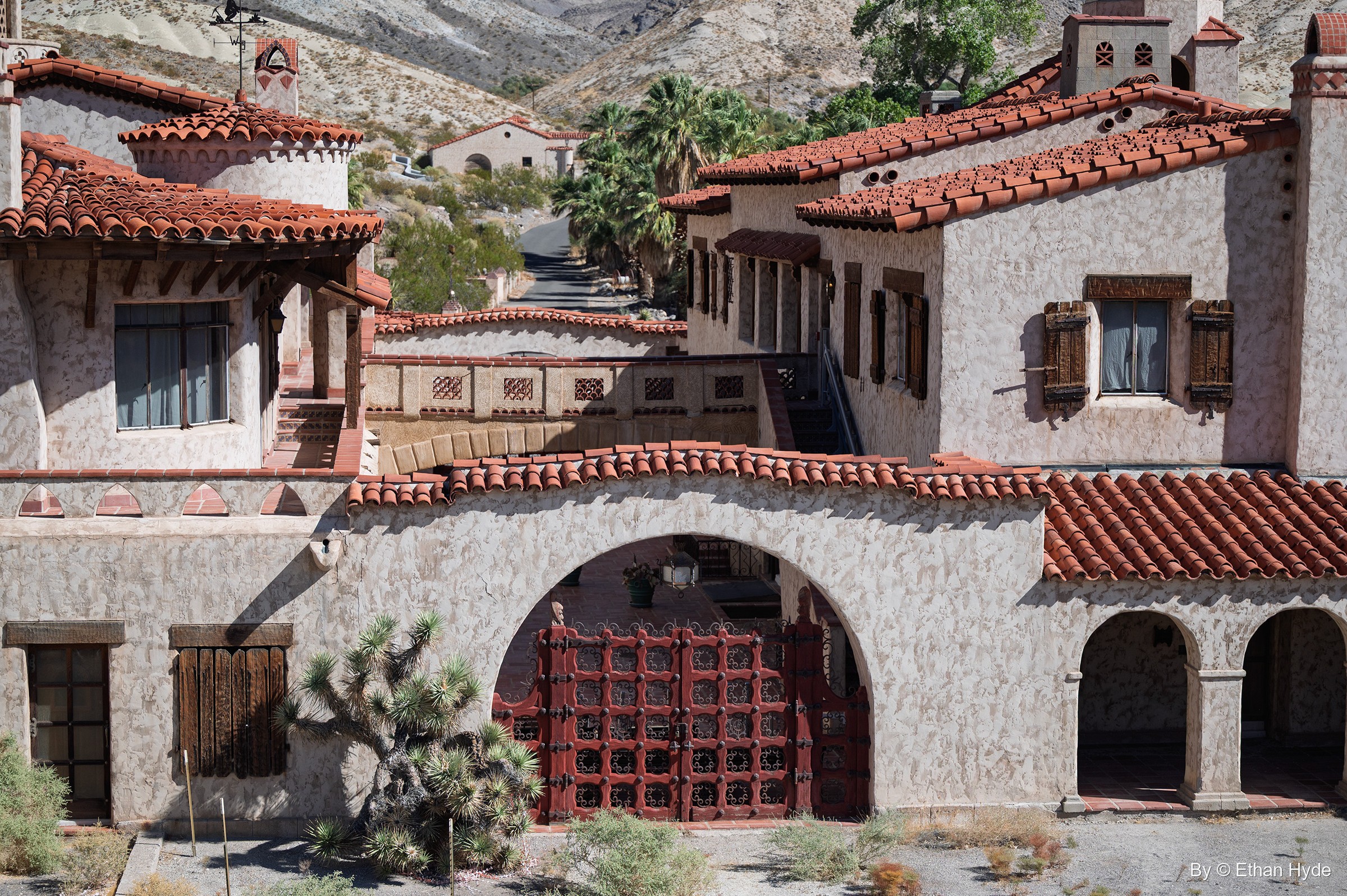 Stucco facade of Scotty's Castle with a red-gated archway and a Joshua tree, rooftops stepping back toward the canyon.