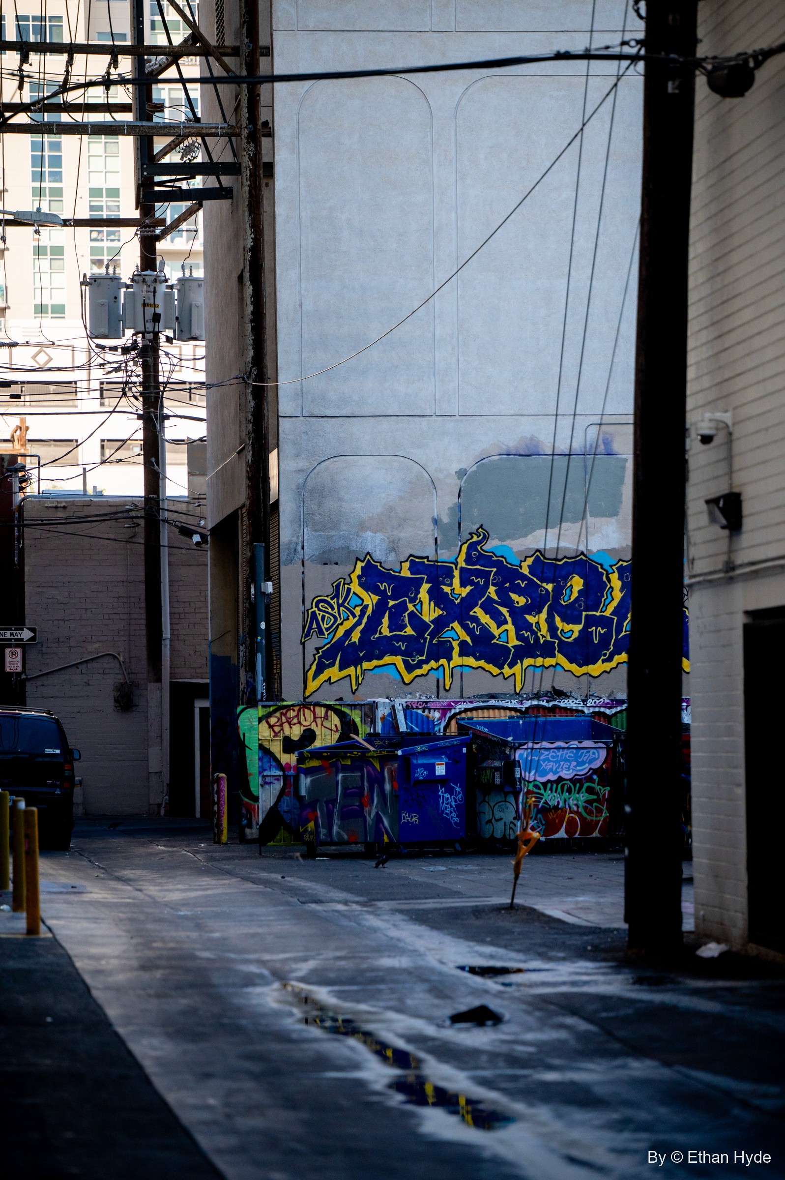 A narrow alley framed by power poles opens onto a blue and yellow graffiti piece over tagged dumpsters.