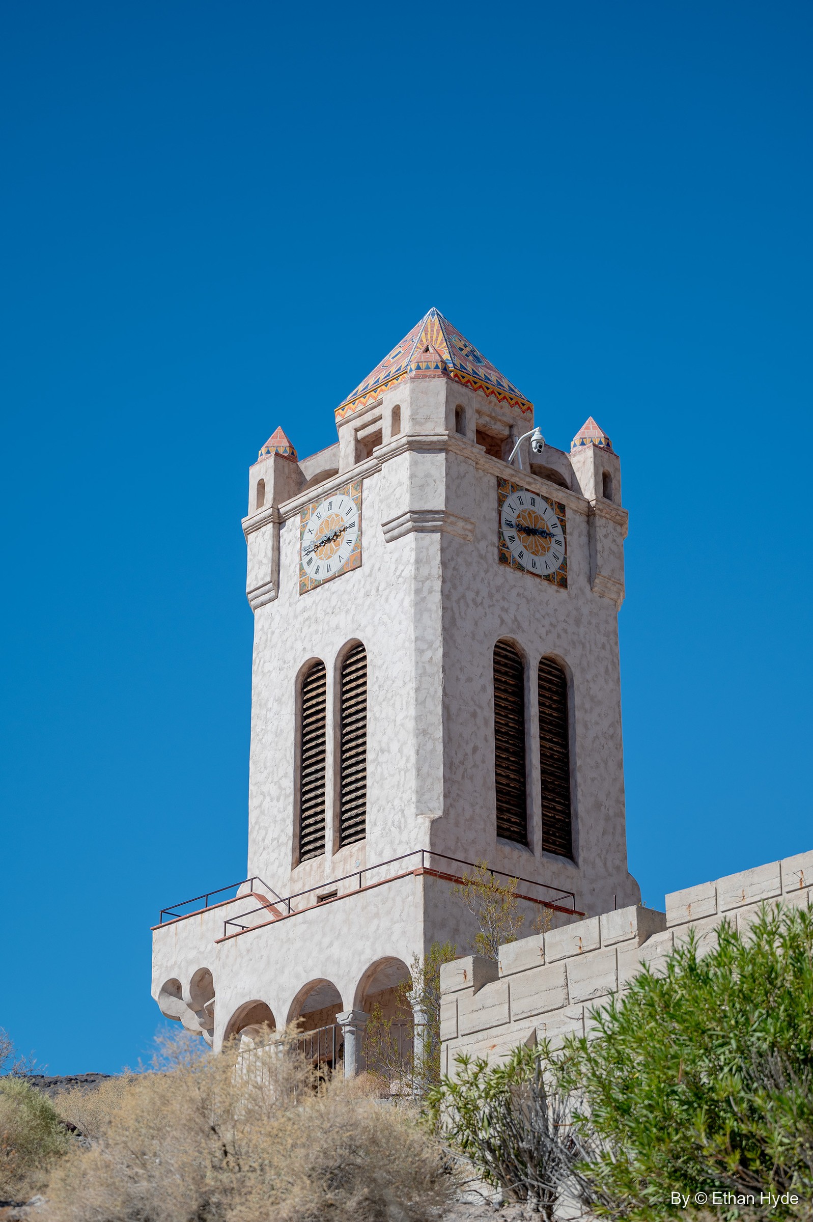 Looking up at the Chimes Tower at Scotty's Castle: clock faces, louvered belfry openings and an arcaded base against a deep blue sky.