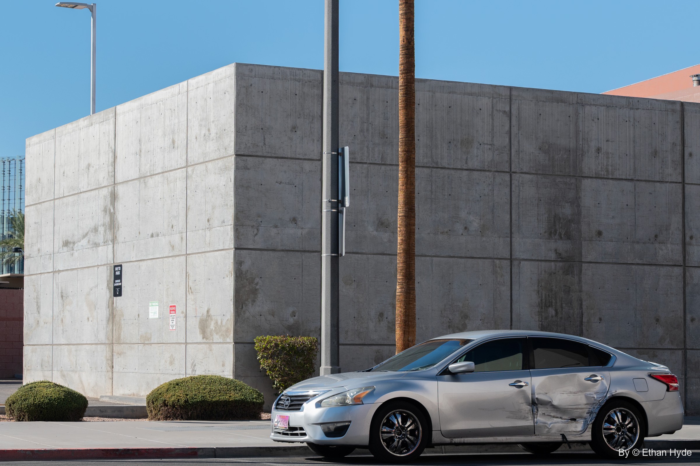 A windowless board-formed concrete building beside palm trunks, a silver sedan with a crumpled door parked in front.