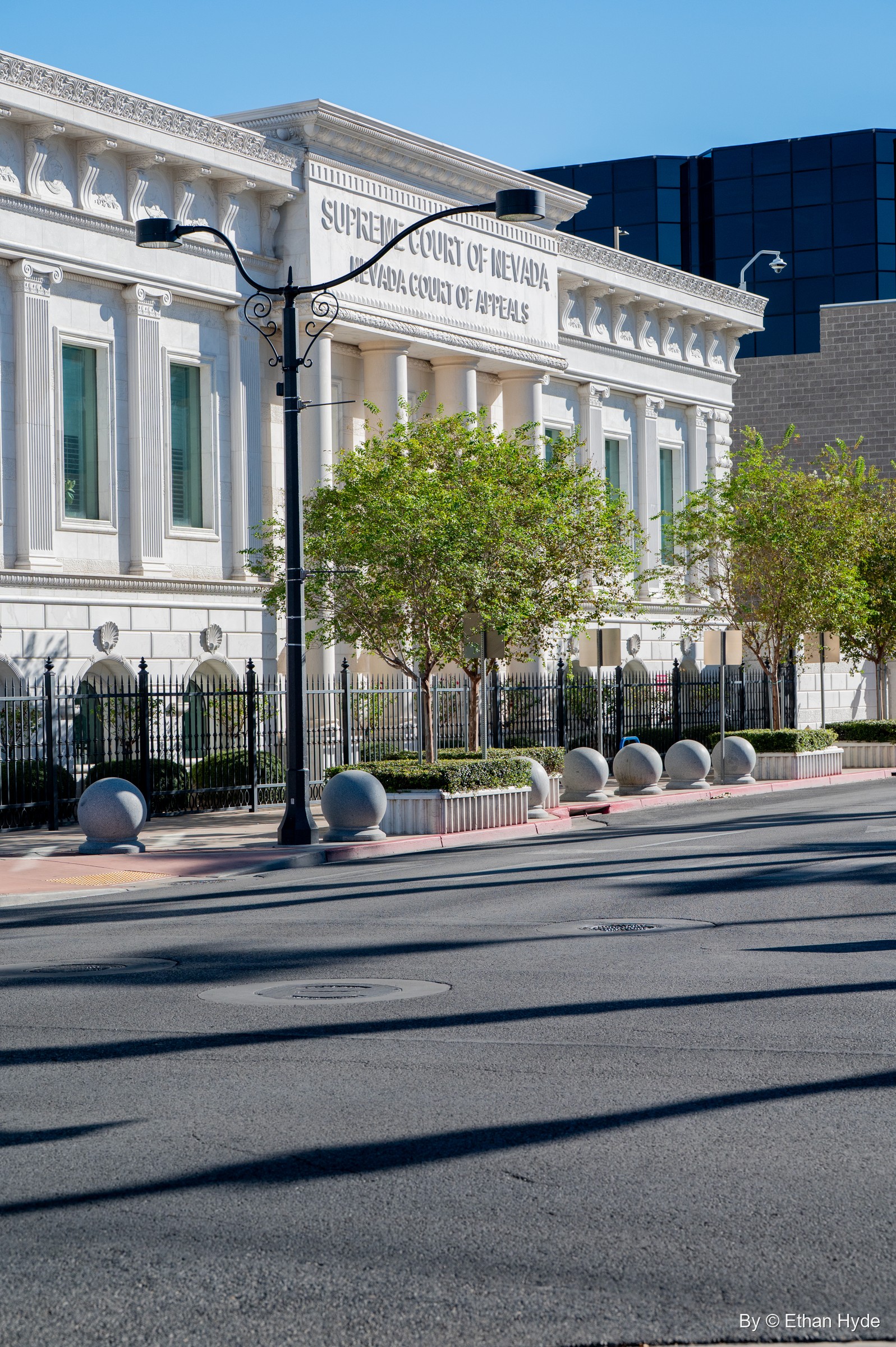 The full white facade of the Nevada Supreme Court building behind young trees and a black street lamp, seen across an empty street.