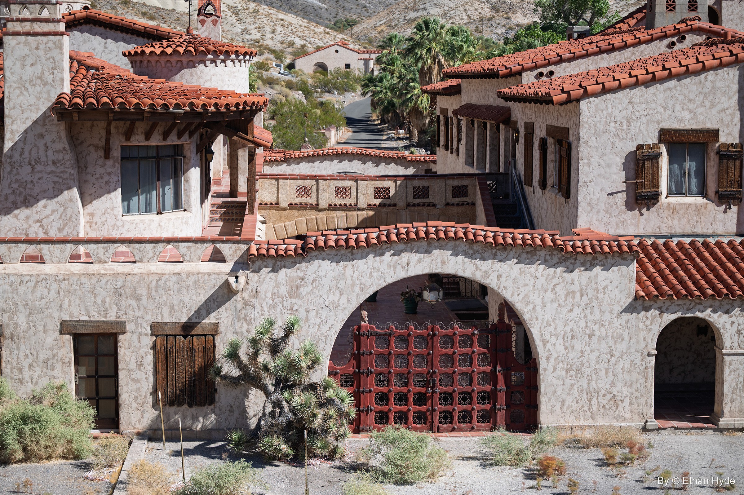 The arched entrance of Scotty's Castle with red wood-and-iron gates, stucco walls and a Joshua tree, beneath tiled rooftops.