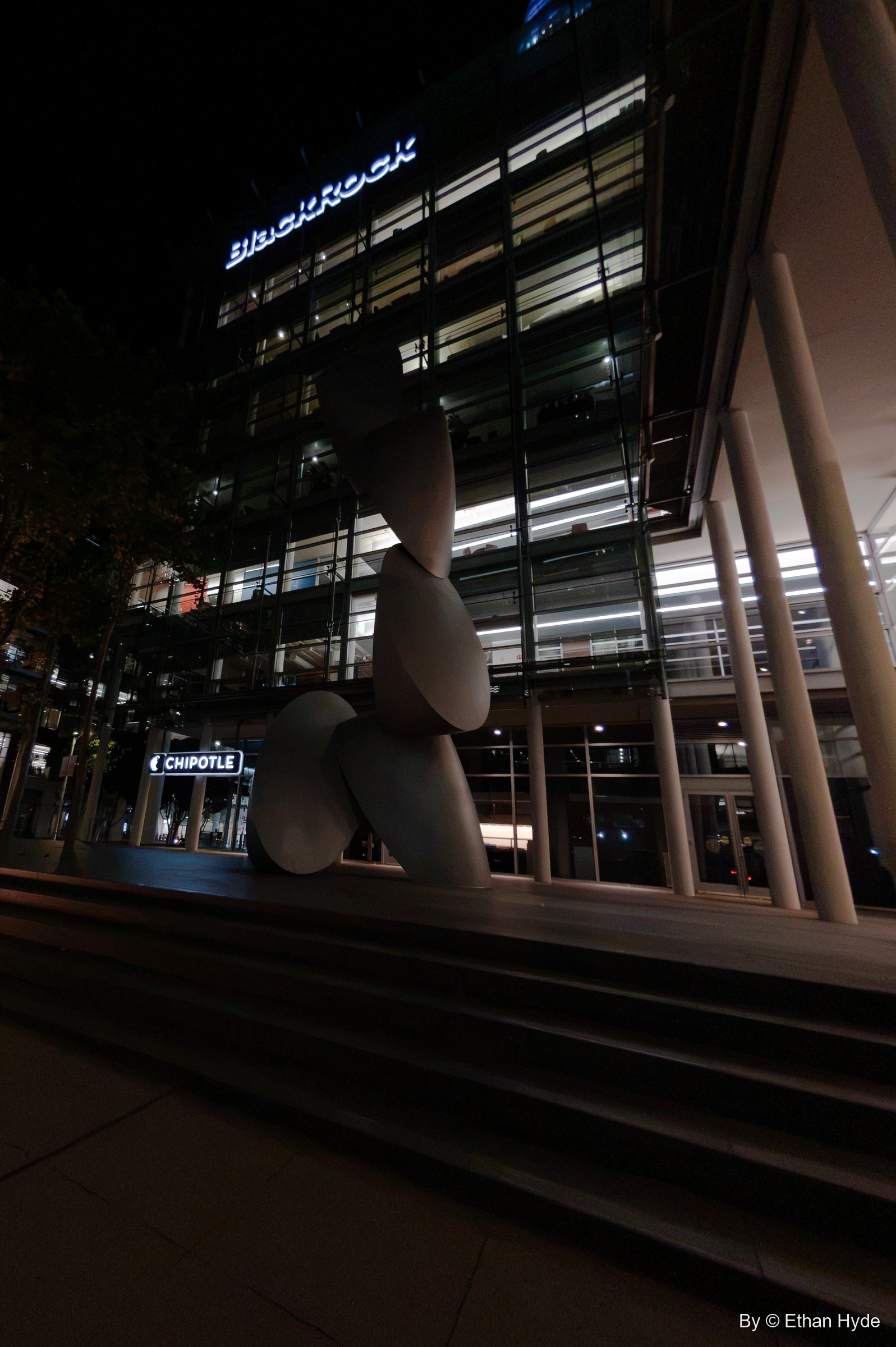 Close vertical view of the stacked disc sculpture Time Signature in front of a lit office facade at night.
