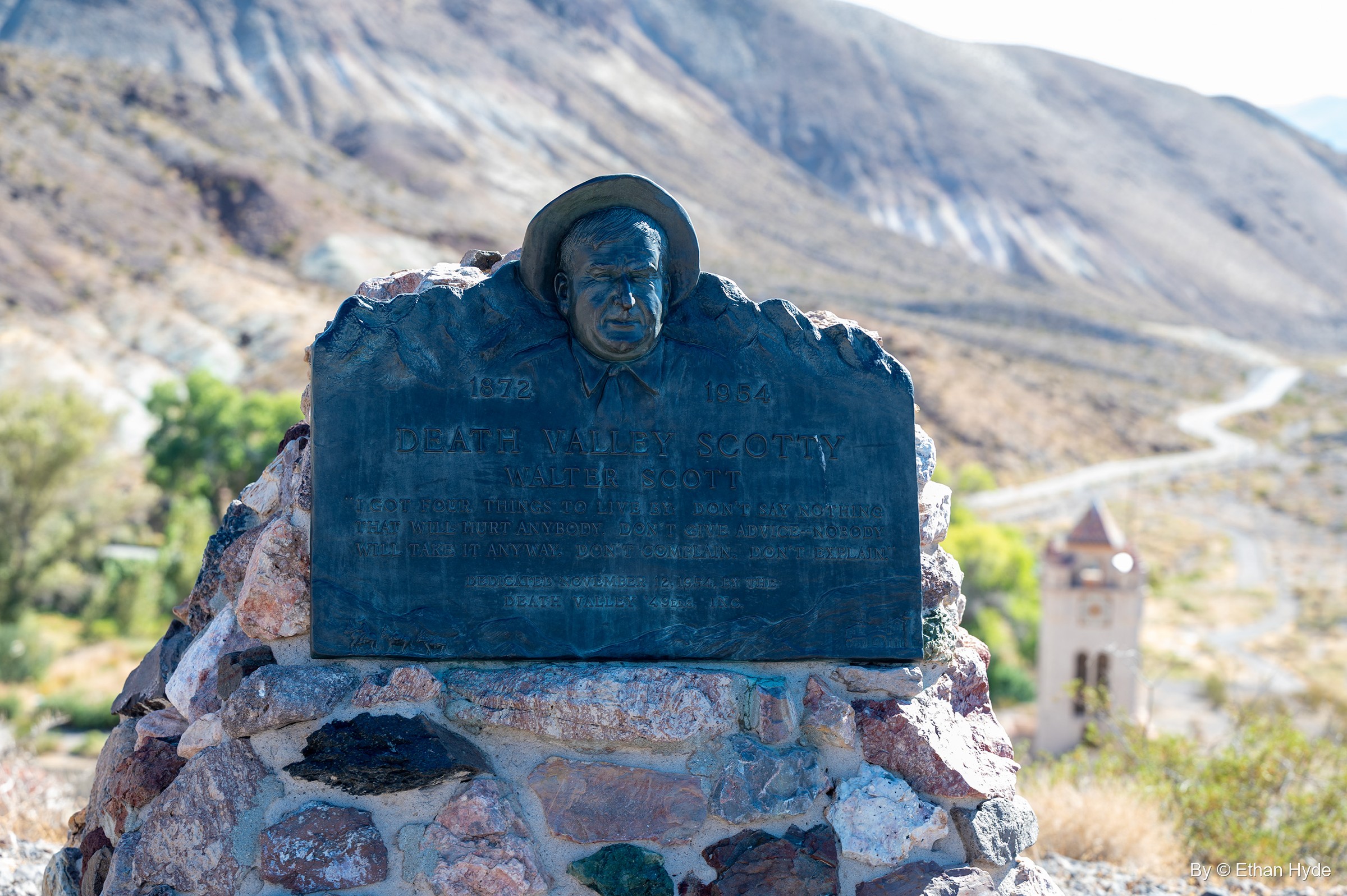 A bronze plaque with a relief portrait of Death Valley Scotty in his hat, set on a stone cairn on Windy Hill, the Chimes Tower and canyon road below.