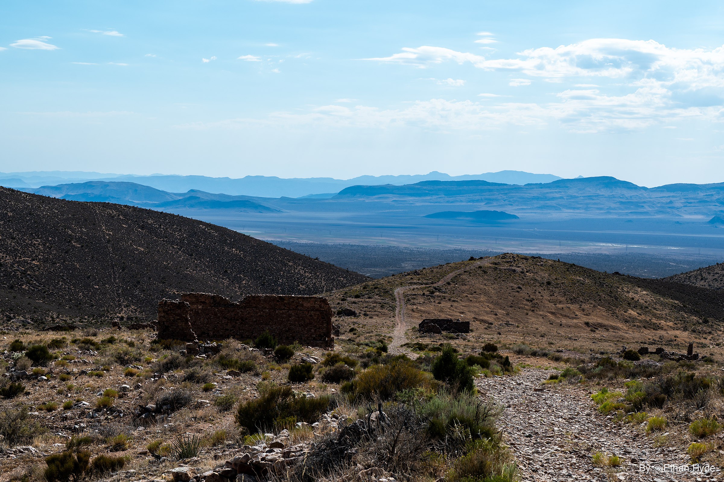 Roofless stone walls of Delamar ruins sit on a scrubby hillside beside a rocky track, with a wide valley and hazy blue ranges beyond.