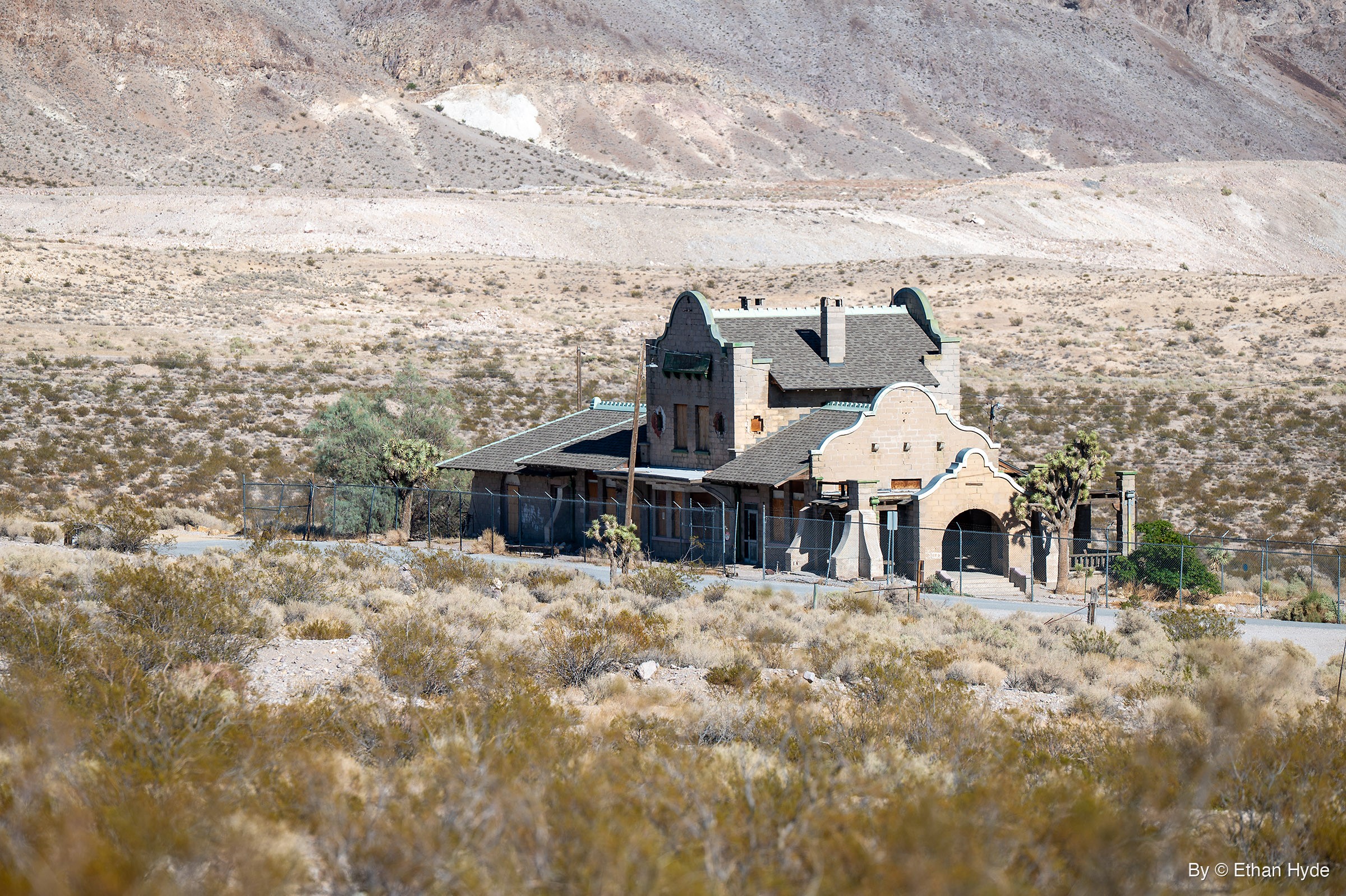 A tan block depot with curved parapet gables, a chimney and a gray shingle roof sits behind a fence among Joshua trees, a bare slope behind.