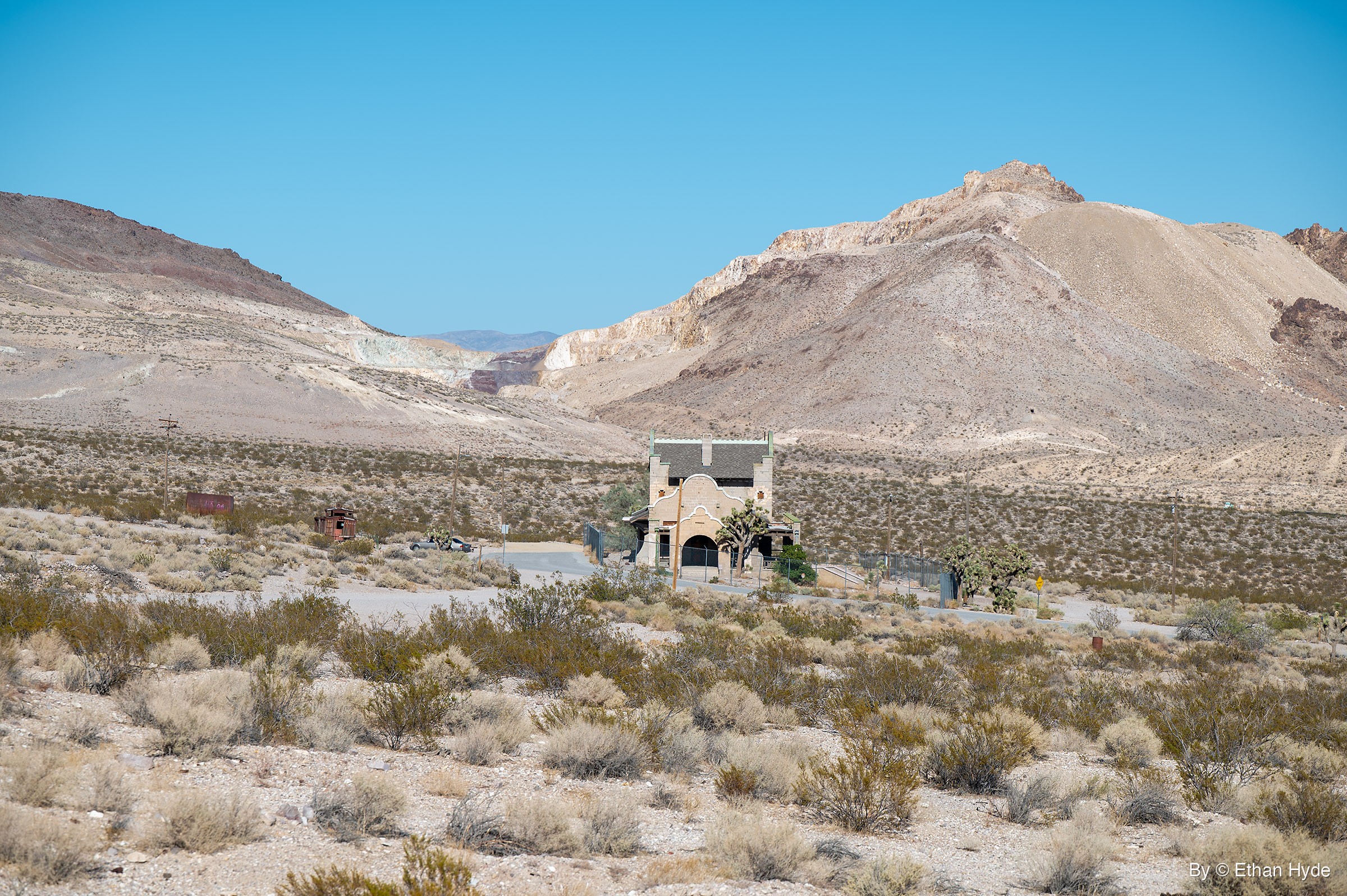 A small mission-style depot sits at the foot of large pale hills, a rusted red caboose at left and desert scrub in the foreground.