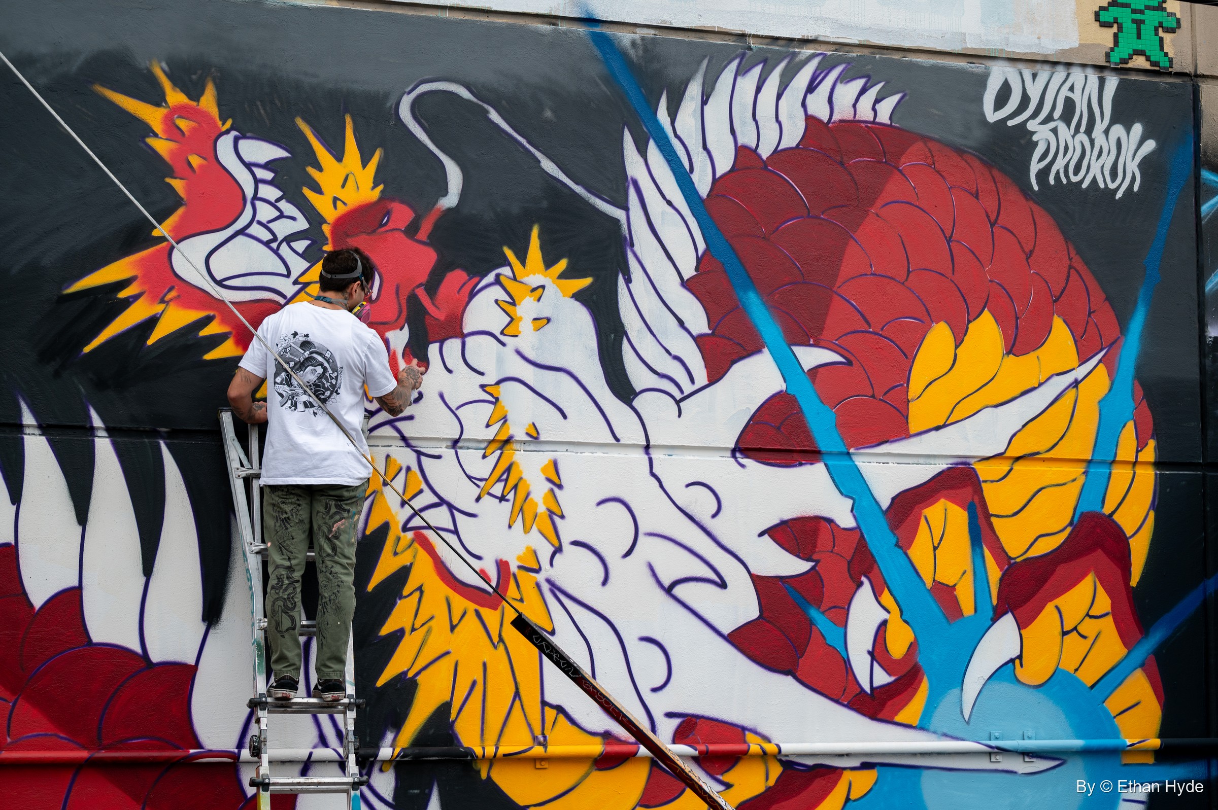 A painter on a stepladder works on a red and gold dragon with a white winged creature, a green pixel-tile figure on the wall above.