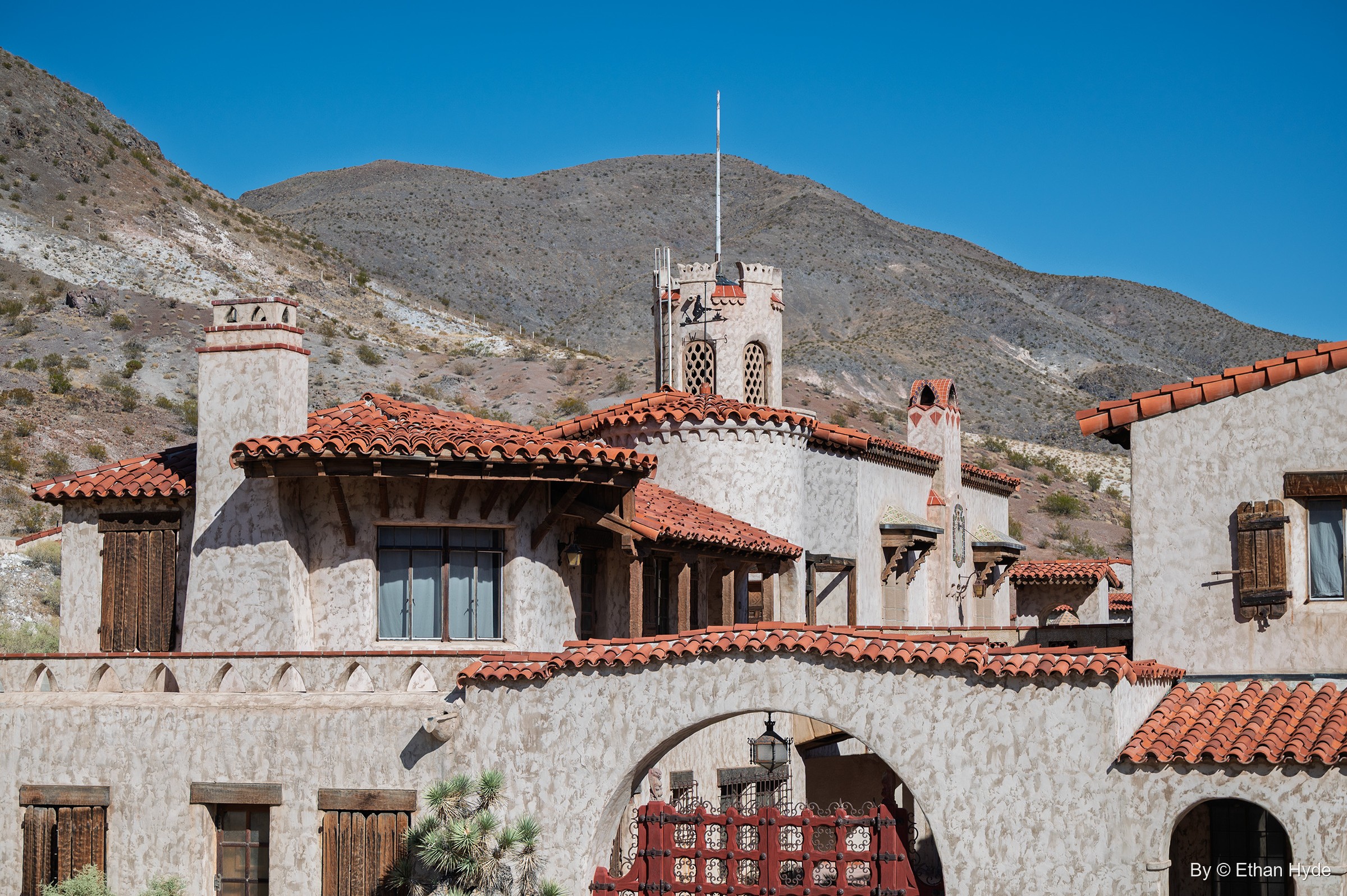 The stucco facade of Scotty's Castle, with tile roofs, chimneys and a crenellated tower beneath a dark desert hill.