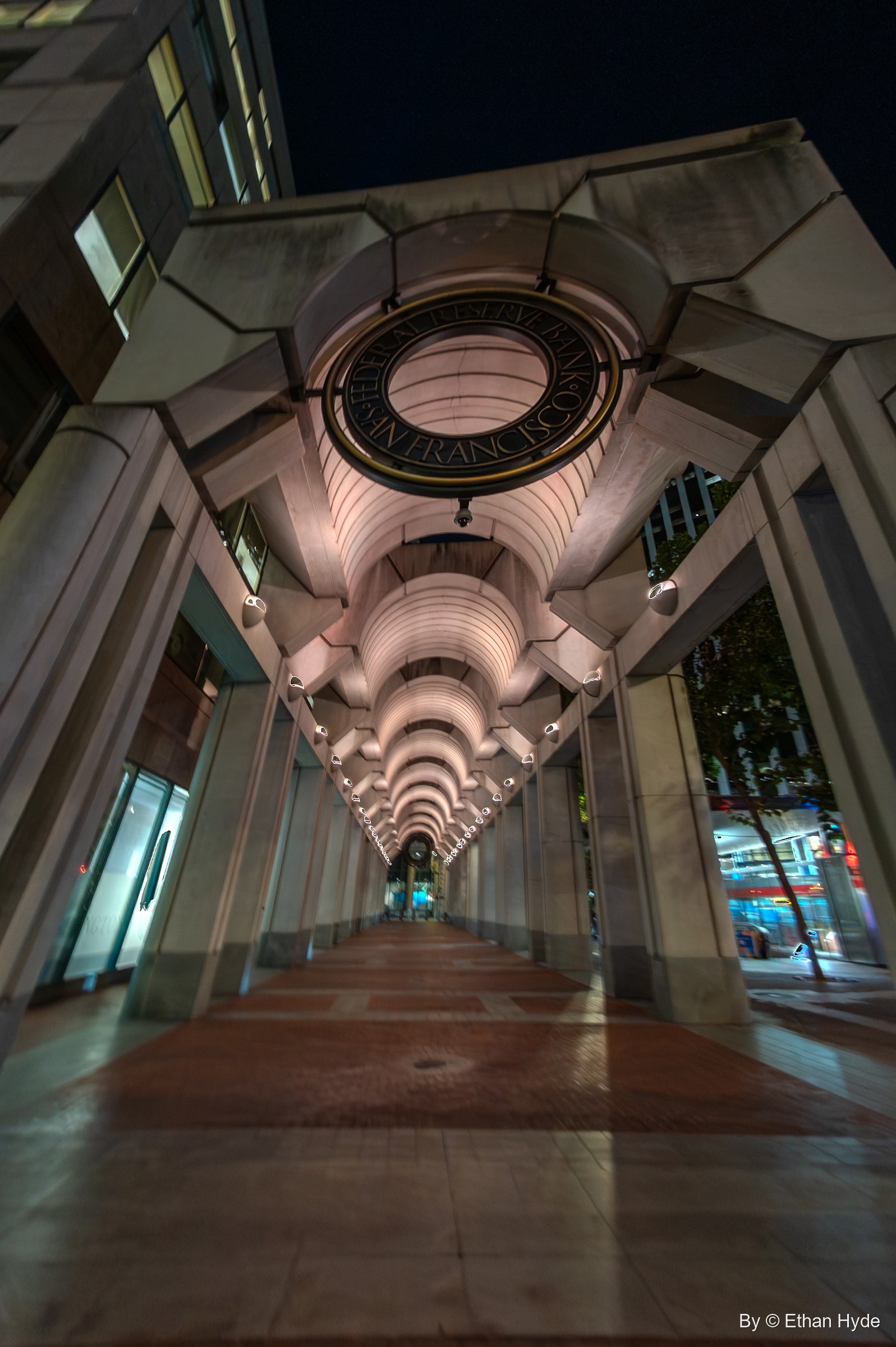 Looking down a vaulted, lit arcade at night beneath a bronze ring reading "Federal Reserve Bank of San Francisco."
