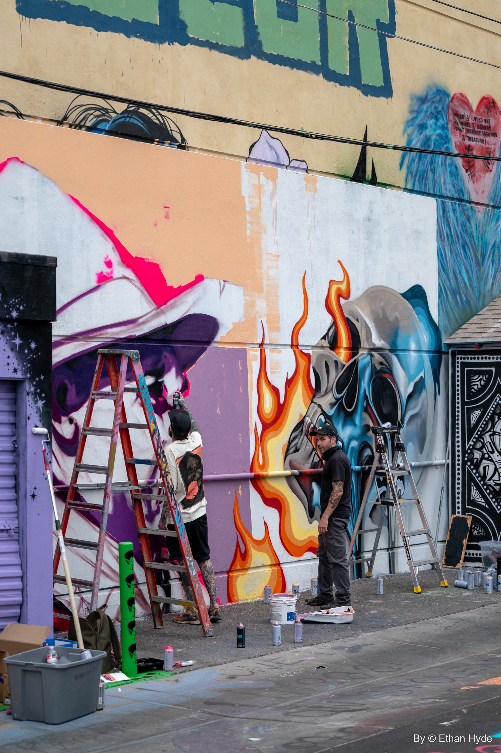 Two painters on ladders work on a mural of flames and a blue skull against a purple and peach wall.