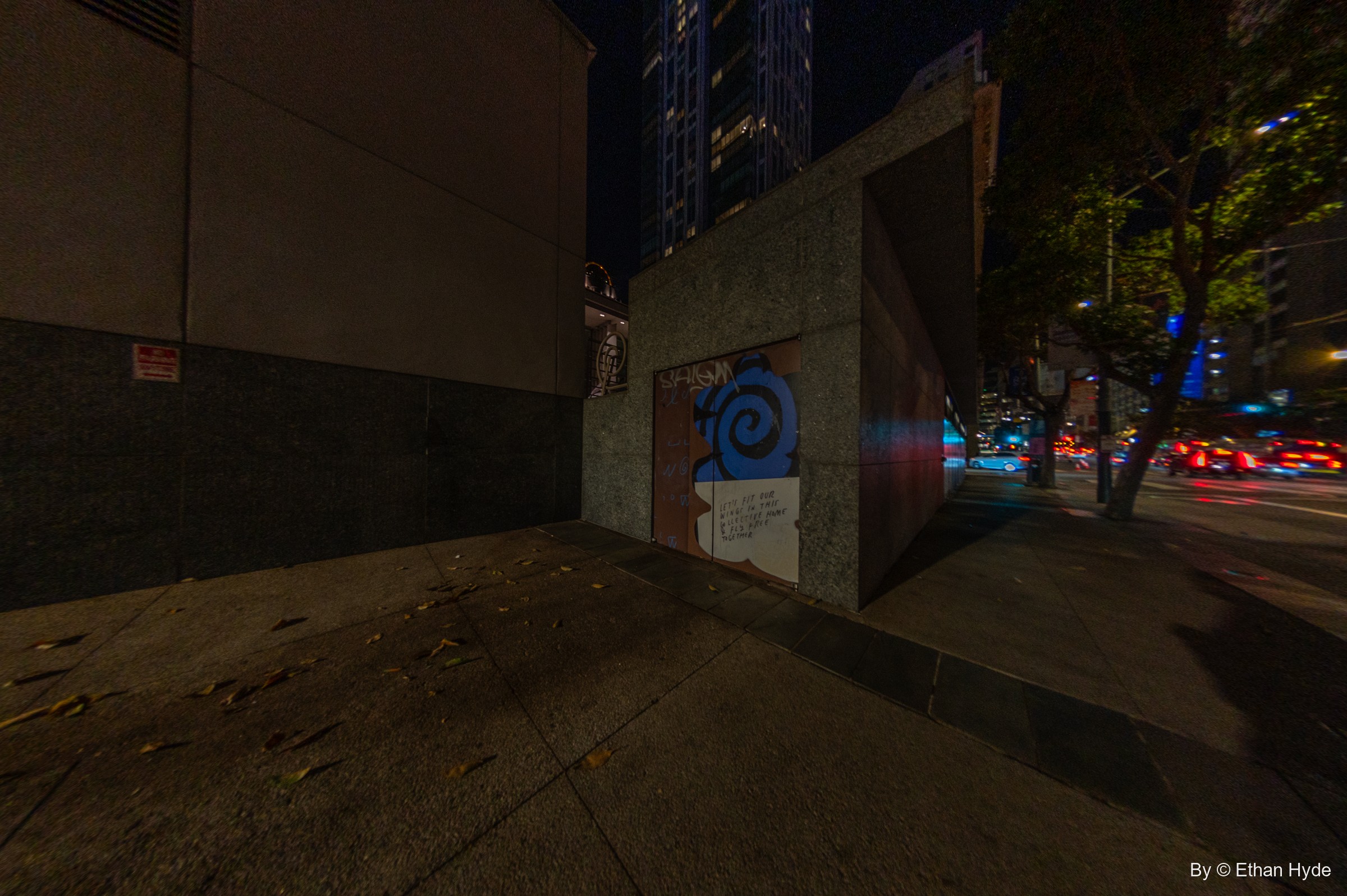 At night, a boarded doorway in a granite wall is painted with a blue spiral and the words "Let's fit our wings in this collective home & fly free together."