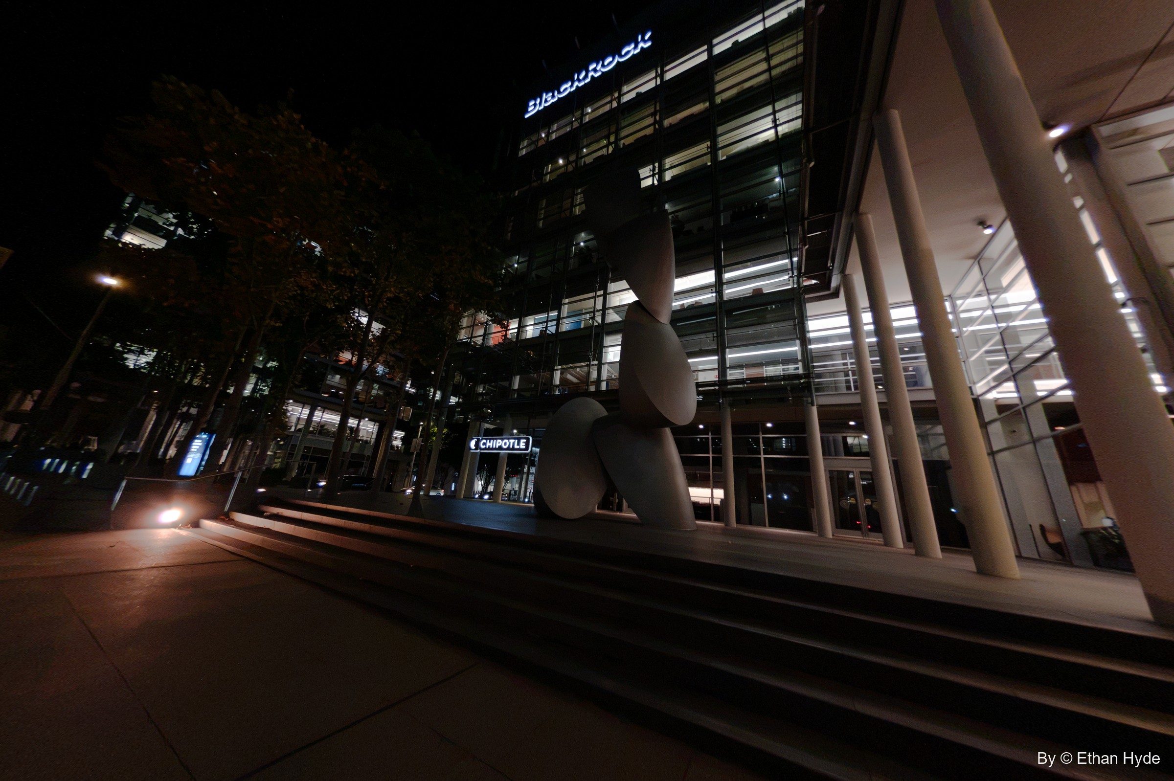 Wide night view of broad steps, angled columns and a stacked white sculpture before a glass office building.