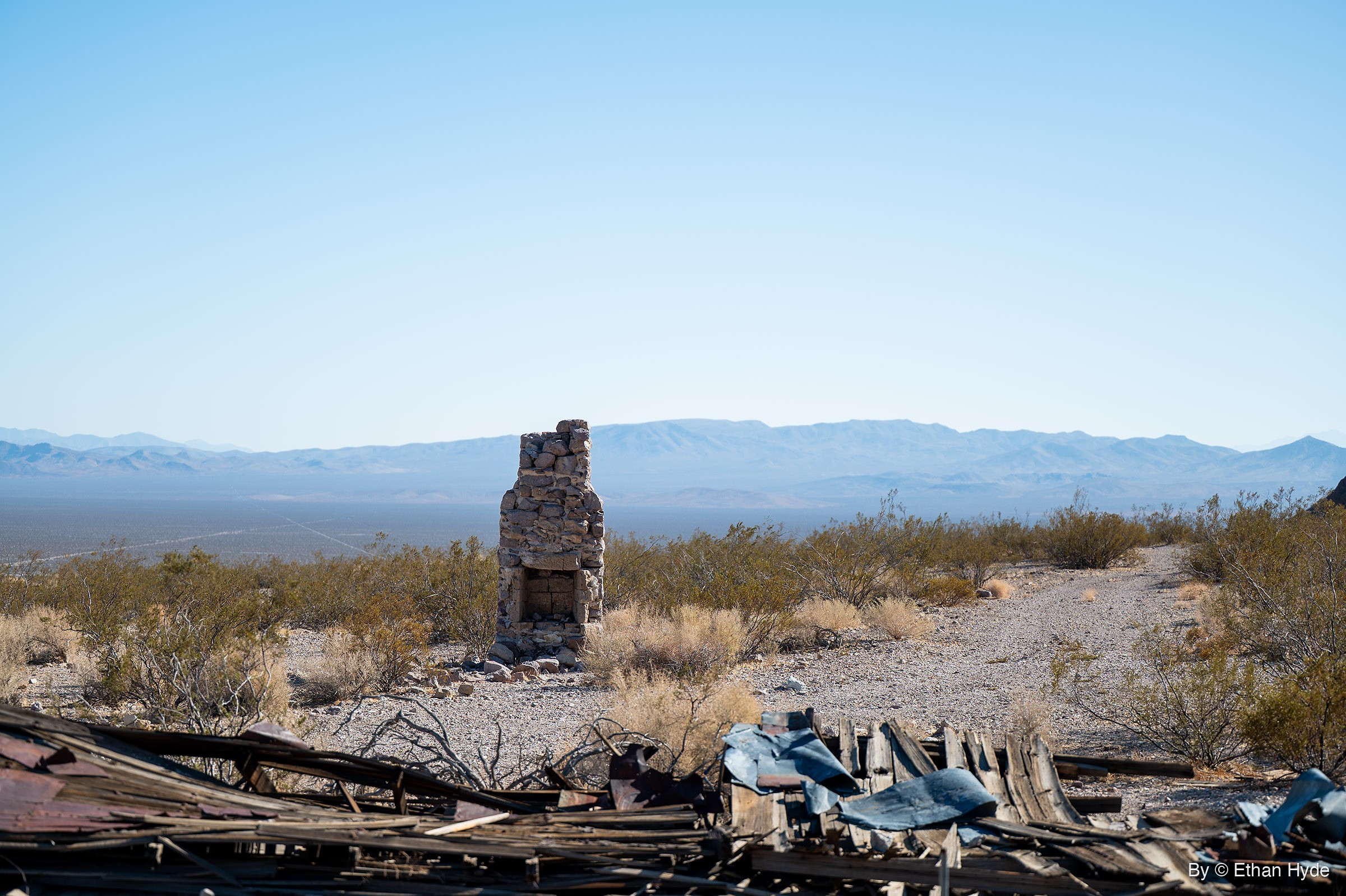 A freestanding fieldstone chimney with an open hearth stands in desert brush, flattened boards and sheet metal in front and a wide valley behind.