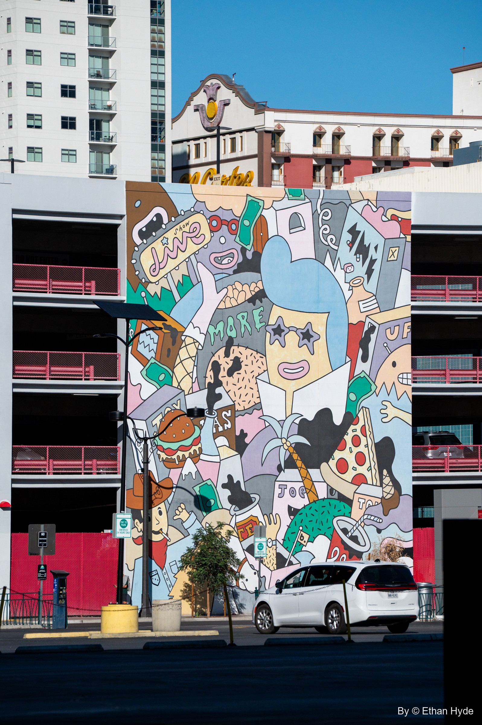 A crowded cartoon mural of faces, food, dice and money covers a parking garage, with an old hotel facade and a tower rising behind it.