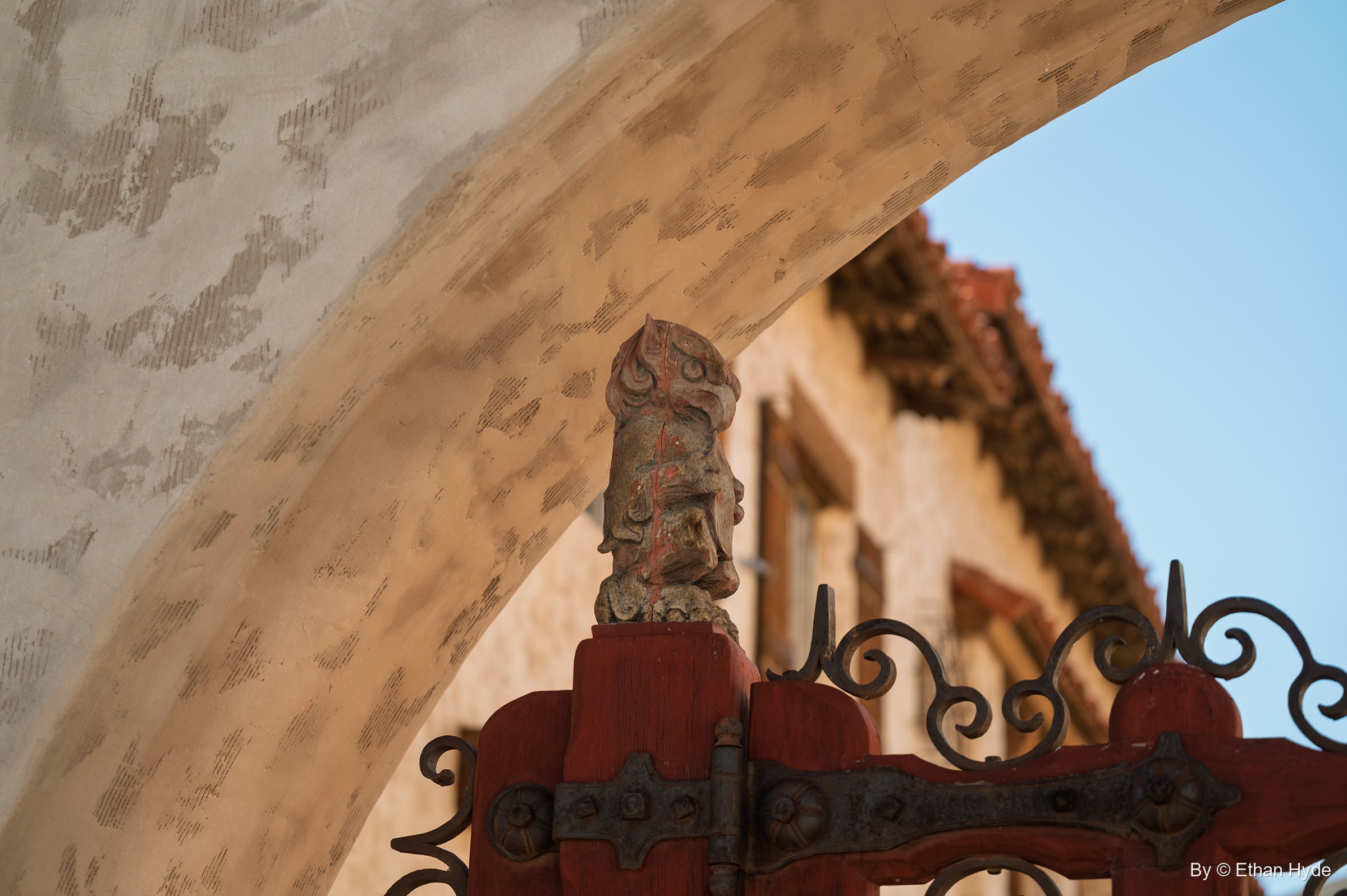 A carved finial atop a red gatepost with an iron strap hinge and scrollwork, framed by a stucco arch at Scotty's Castle.