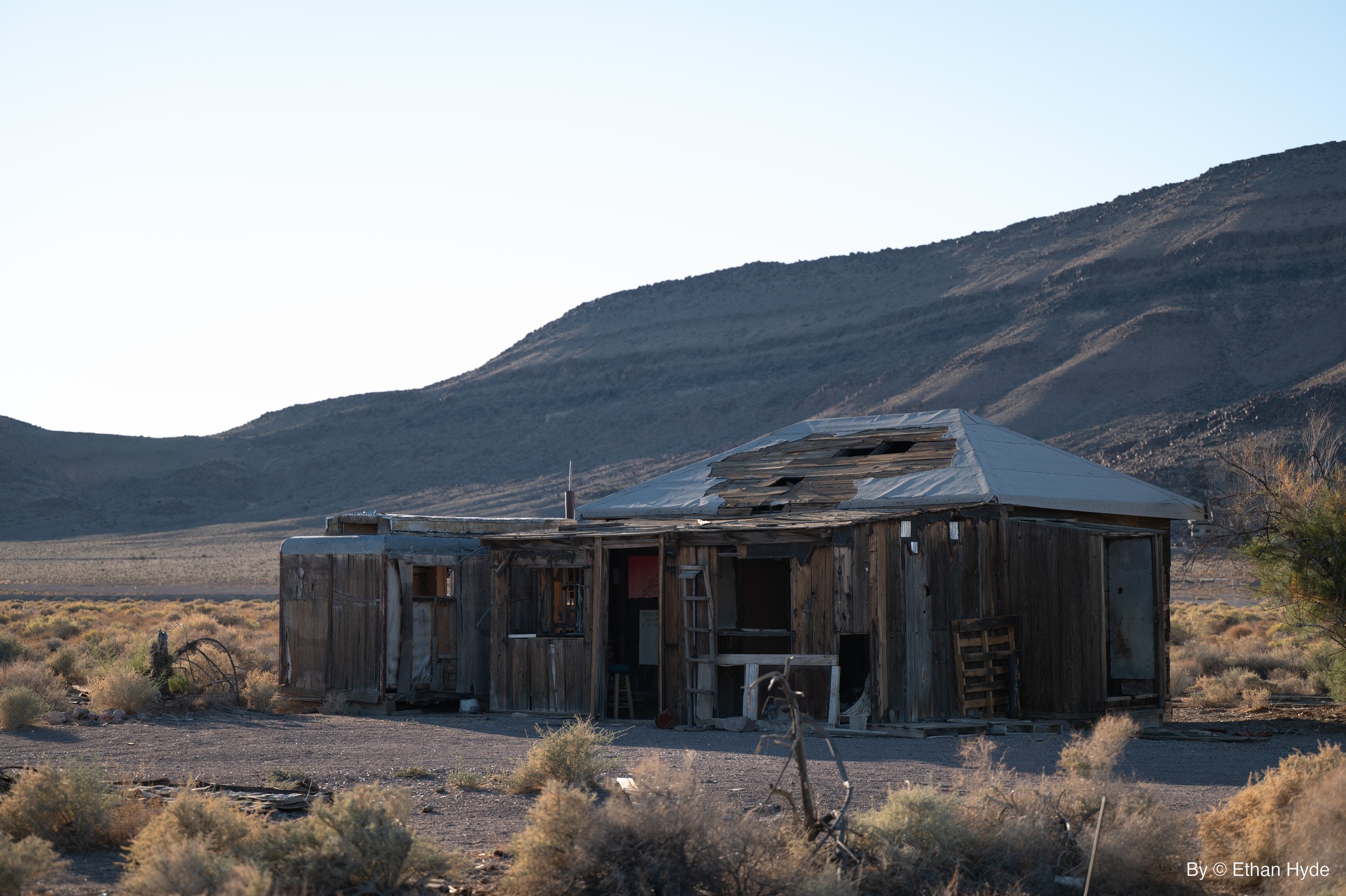A weathered wooden shack standing alone in the desert below distant mountains.