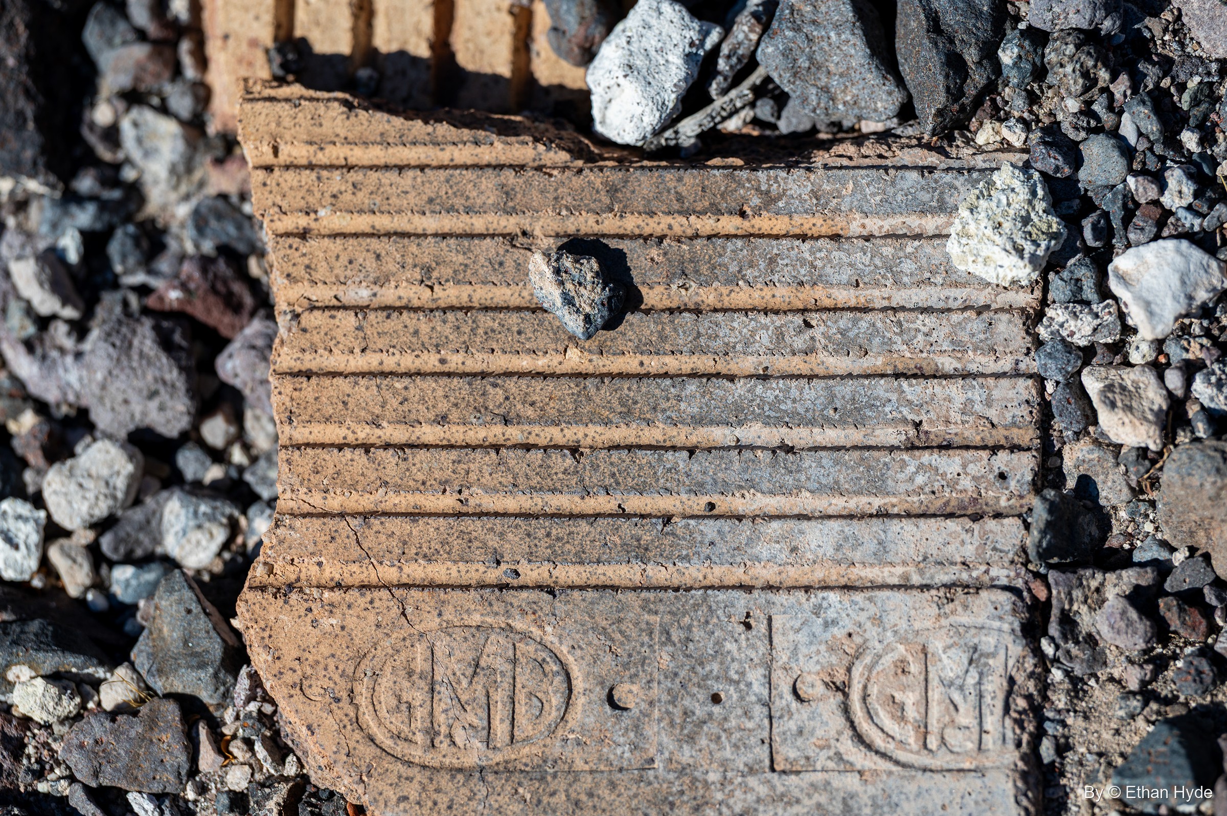 A broken ribbed clay block stamped with an oval "GMB" monogram, lying in volcanic gravel near Scotty's Castle.