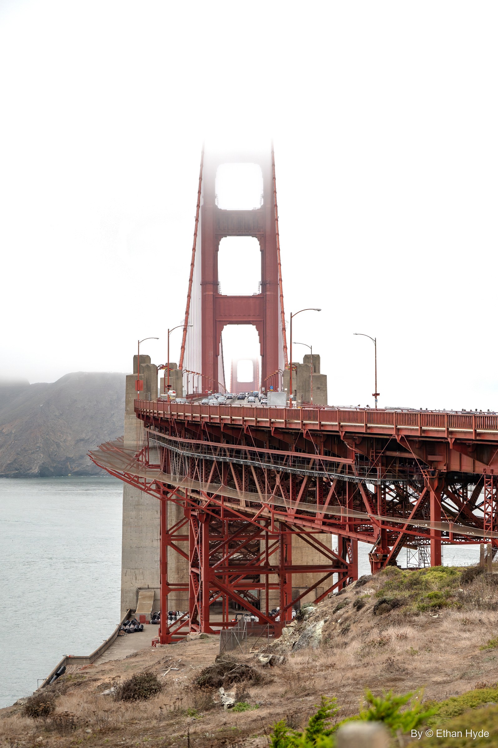 The Golden Gate Bridge's south tower fades into fog above the red steel approach span and a grassy bluff.