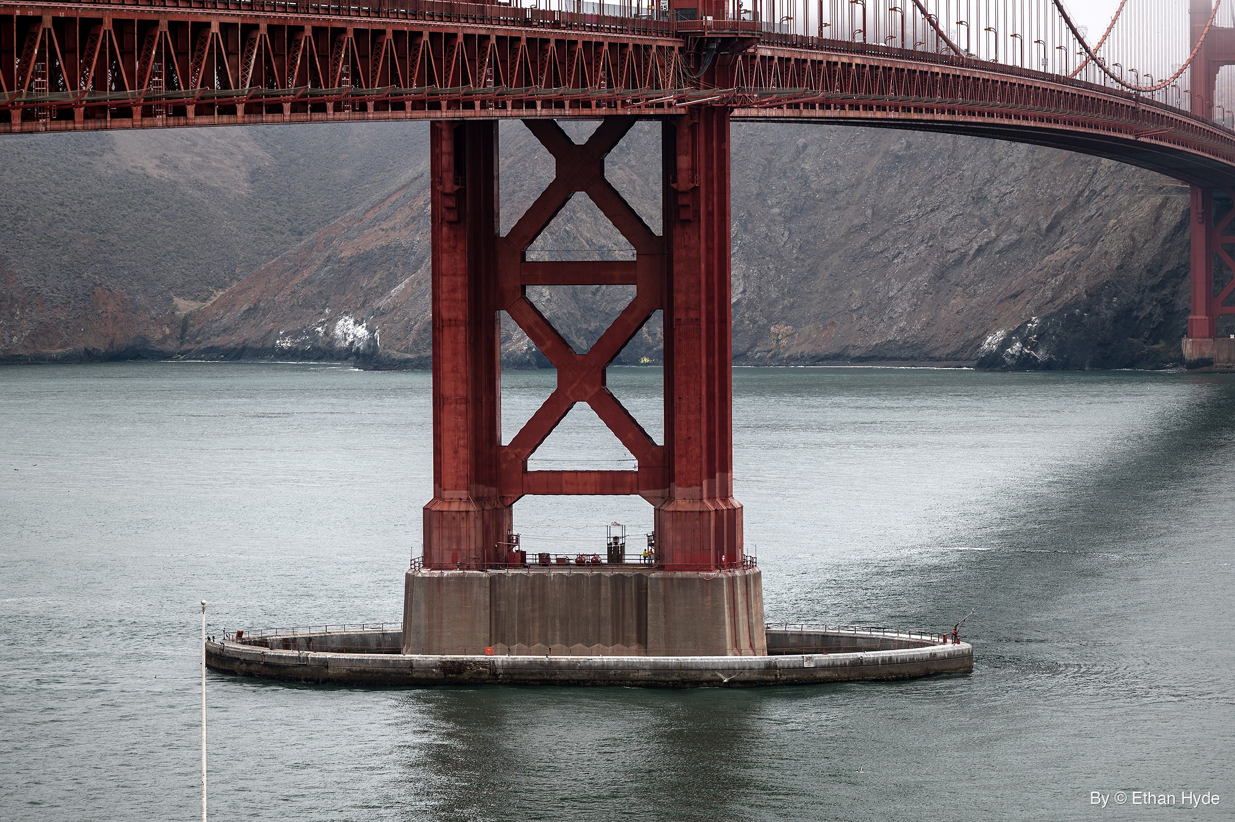 Close view of a red Golden Gate Bridge tower standing on its concrete pier and fender ring in gray-green water, with workers on the pier and rocky headlands behind.