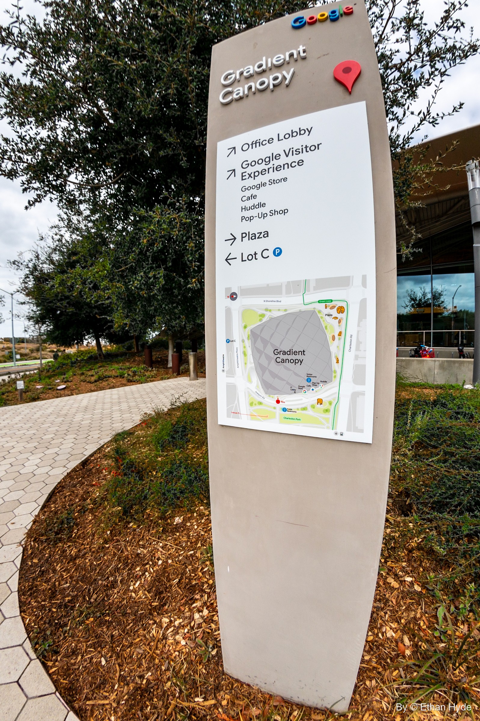 A curved wayfinding sign for Google's Gradient Canopy with a campus map, beside a path and mulched planting.