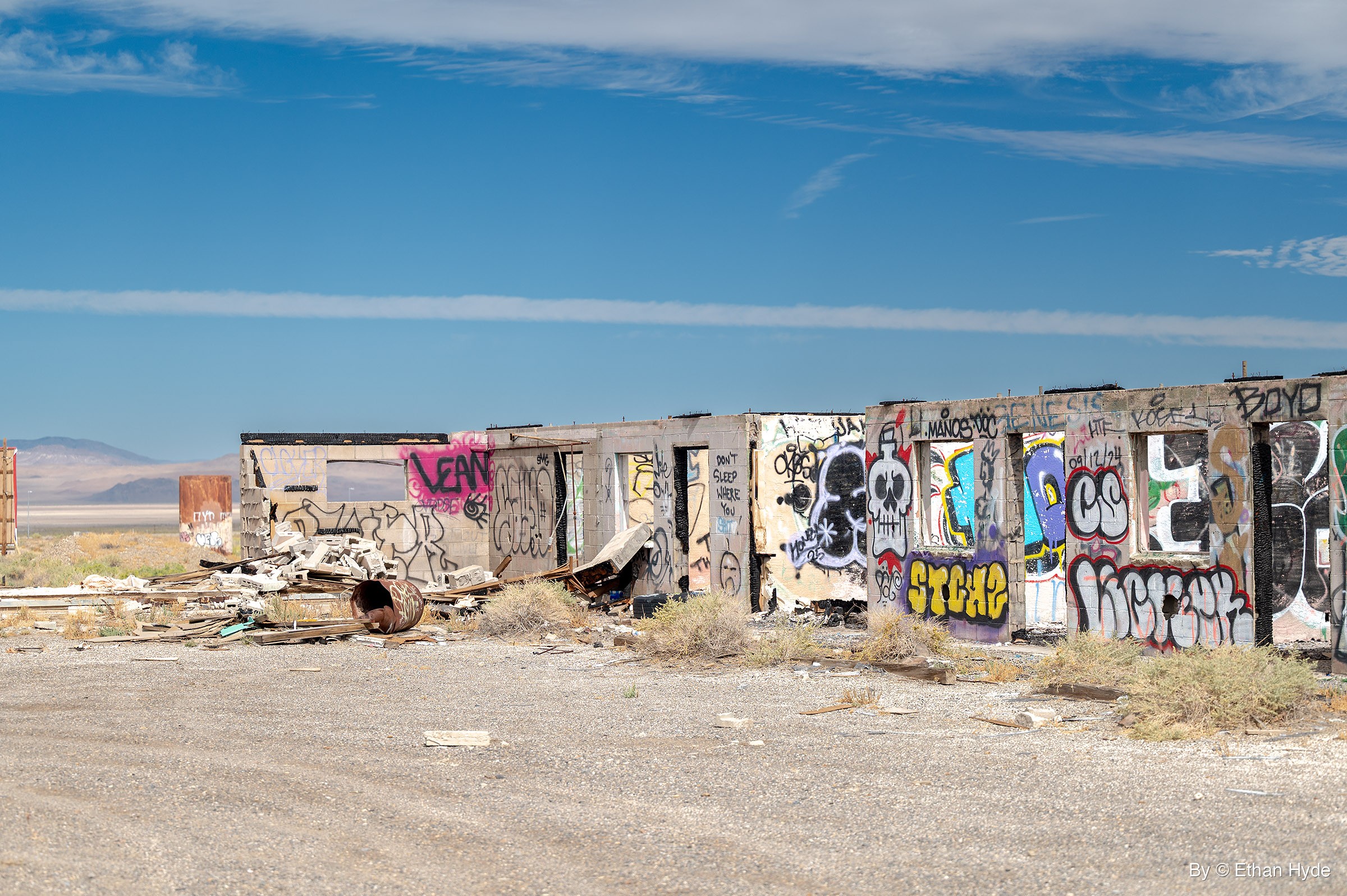 A long row of roofless concrete ruins covered in colorful graffiti, with scattered debris and a rusted barrel in front and a pale basin beyond.