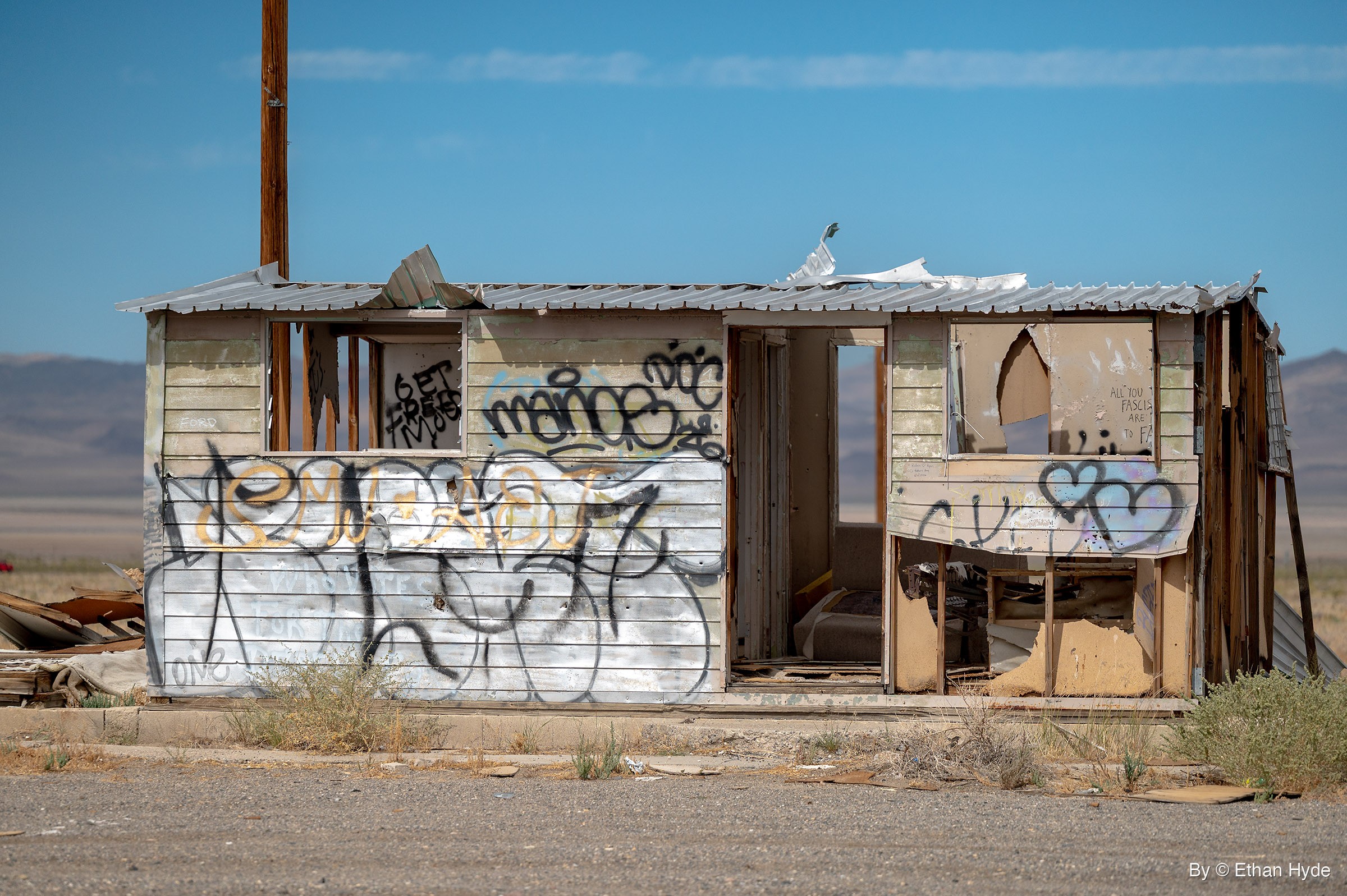A small abandoned shack with a peeling metal roof and graffiti-covered walls, a utility pole behind it and desert mountains on the horizon.