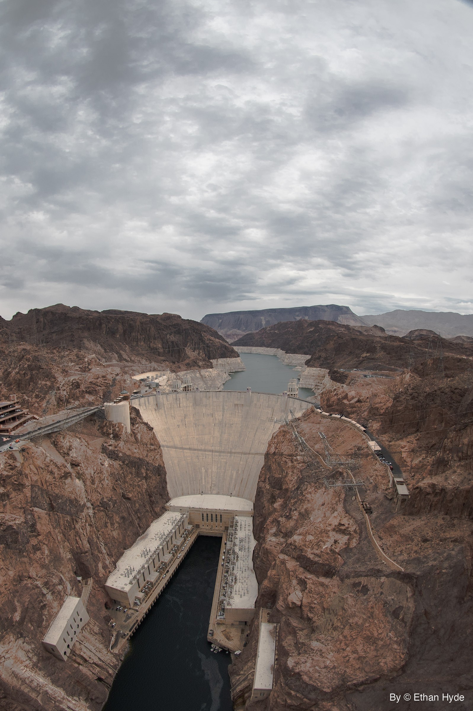 Vertical fisheye view down onto Hoover Dam, its powerhouses and the dark river below, with the reservoir and a mesa behind under heavy cloud.