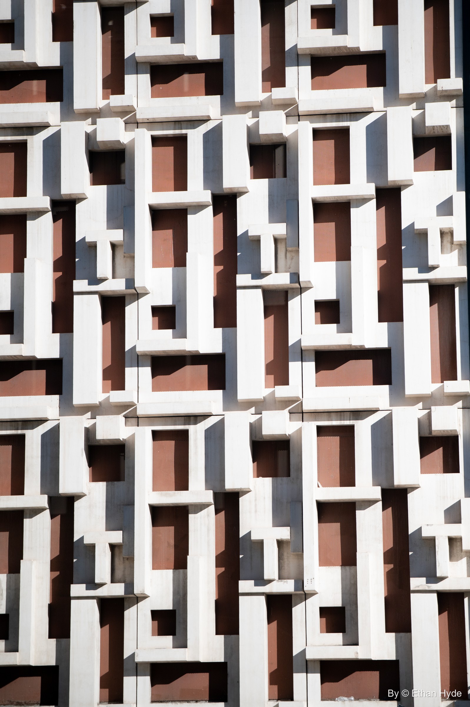 A vertical abstract of a facade of white concrete relief blocks set in a repeating maze-like pattern over brown.