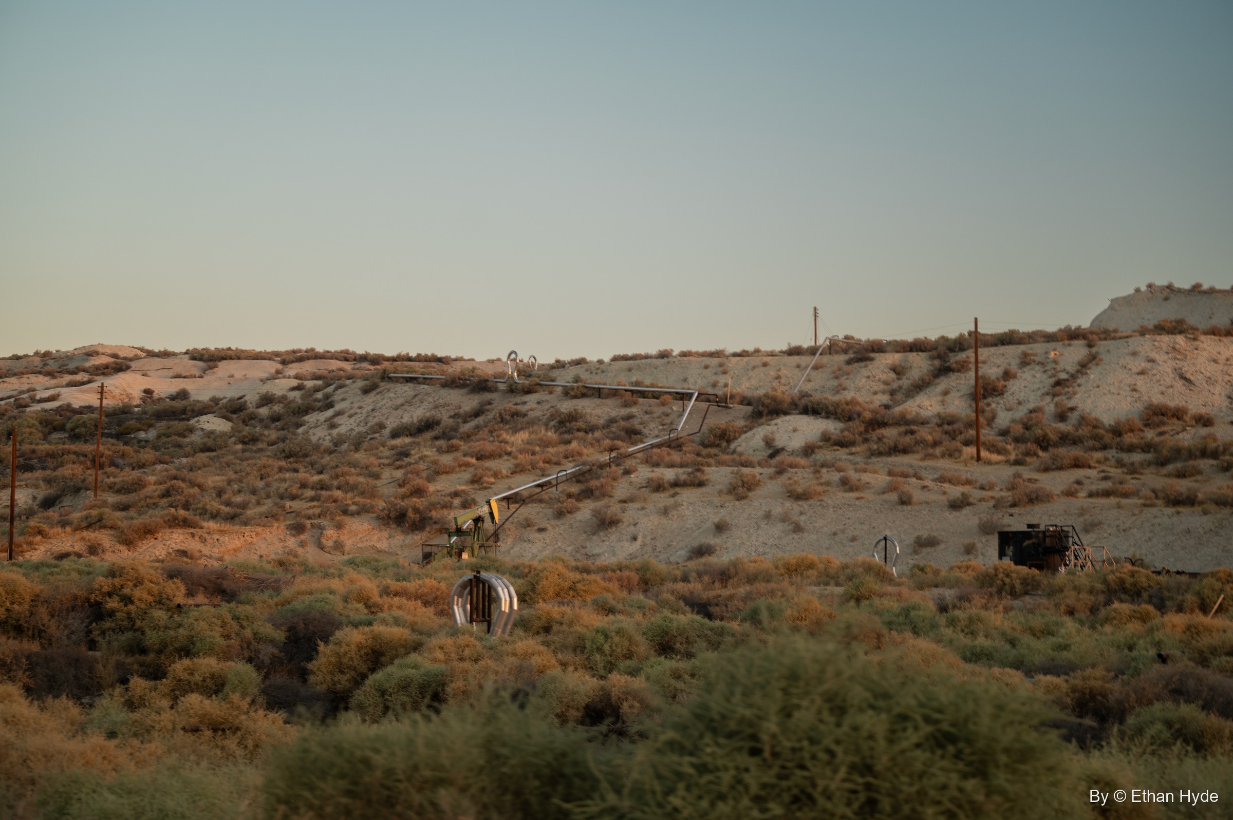 Early light on a scrubby hillside of the McKittrick oil field, scattered with pipes, wellheads and power poles.