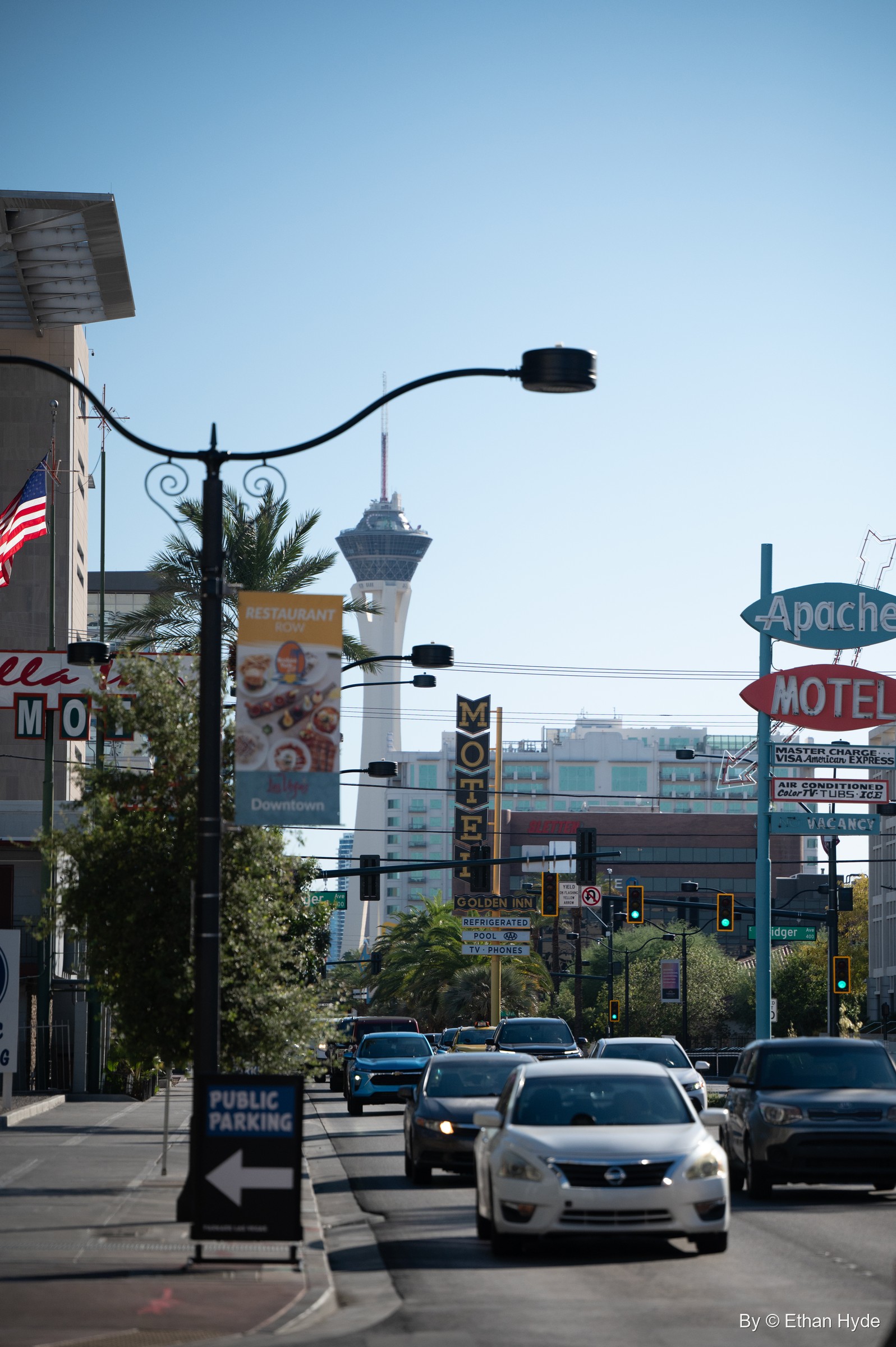 A downtown street of motel signs, palms and street lamps, with a tall observation tower rising in the distance.