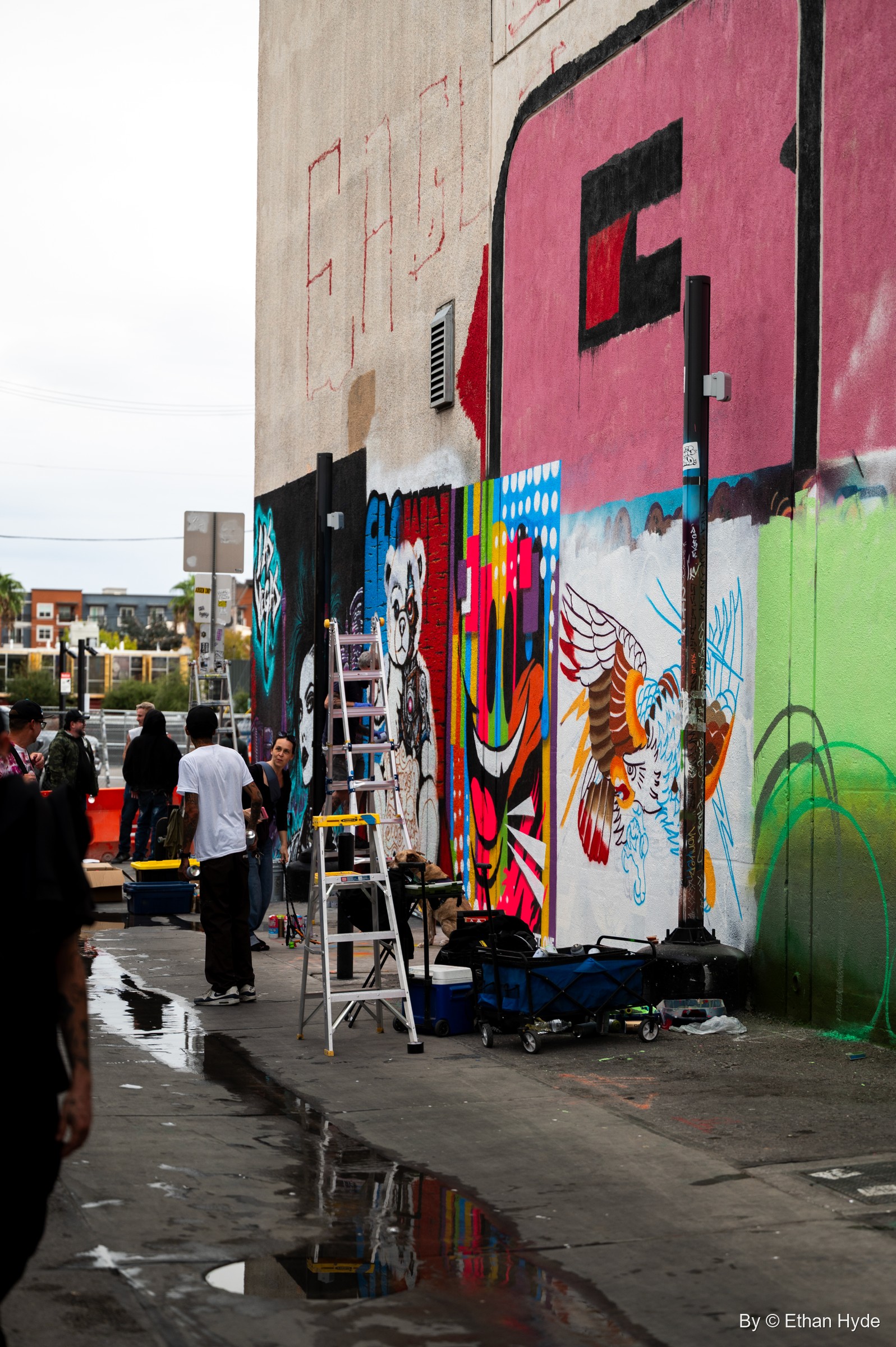 A wet alley lined with half-finished murals, ladders and paint carts, with painters at work and huge pink letters on the wall above.