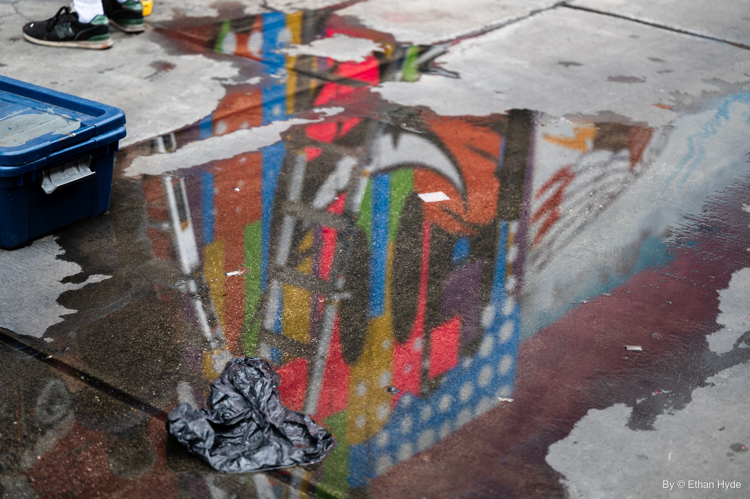 A rain puddle on the sidewalk reflects a brightly colored mural, next to a blue paint tote and a crumpled plastic bag.