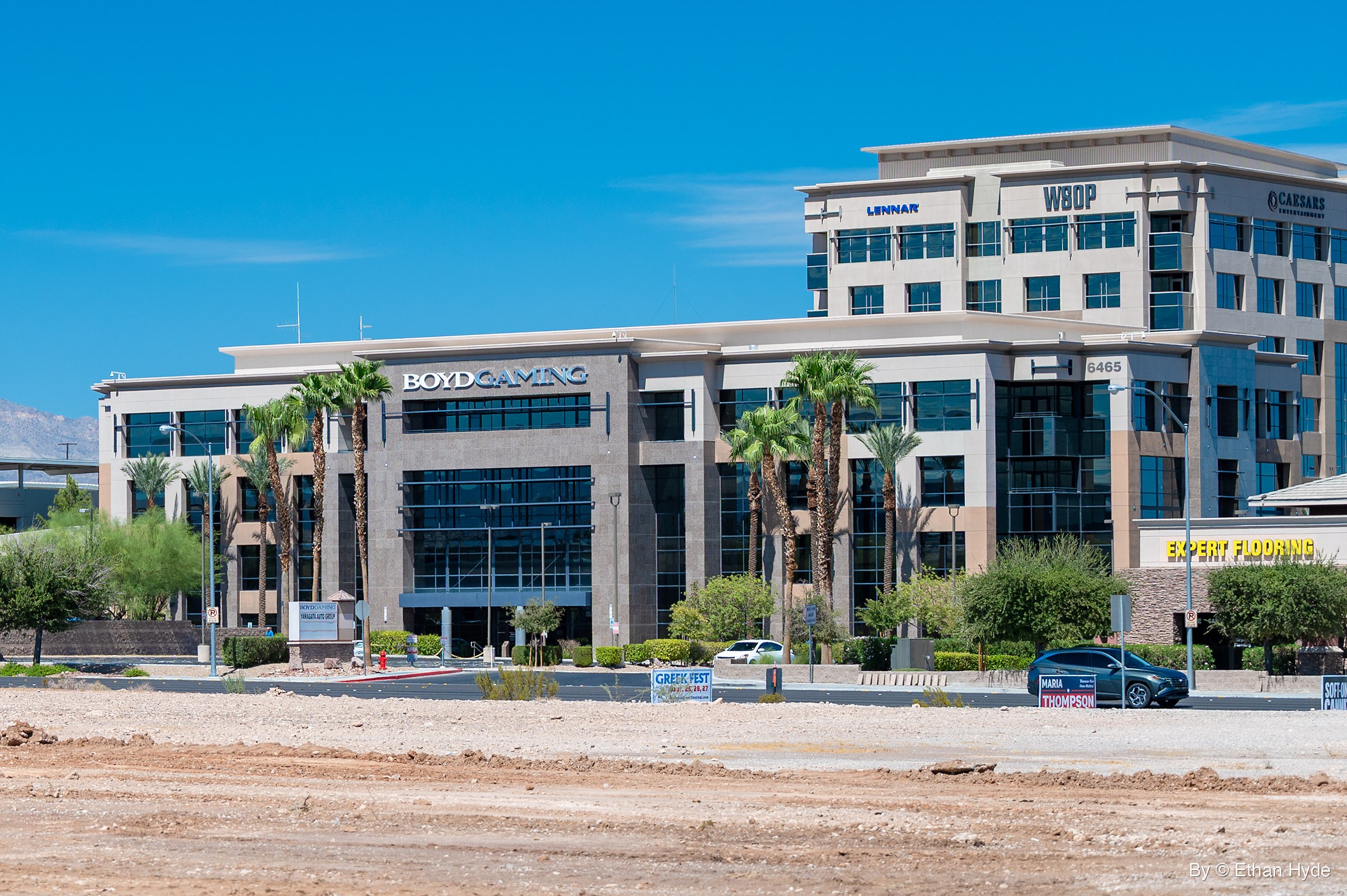 A four-story granite and glass office building with a taller office tower behind it, palm trees along the street and a dirt lot in front.