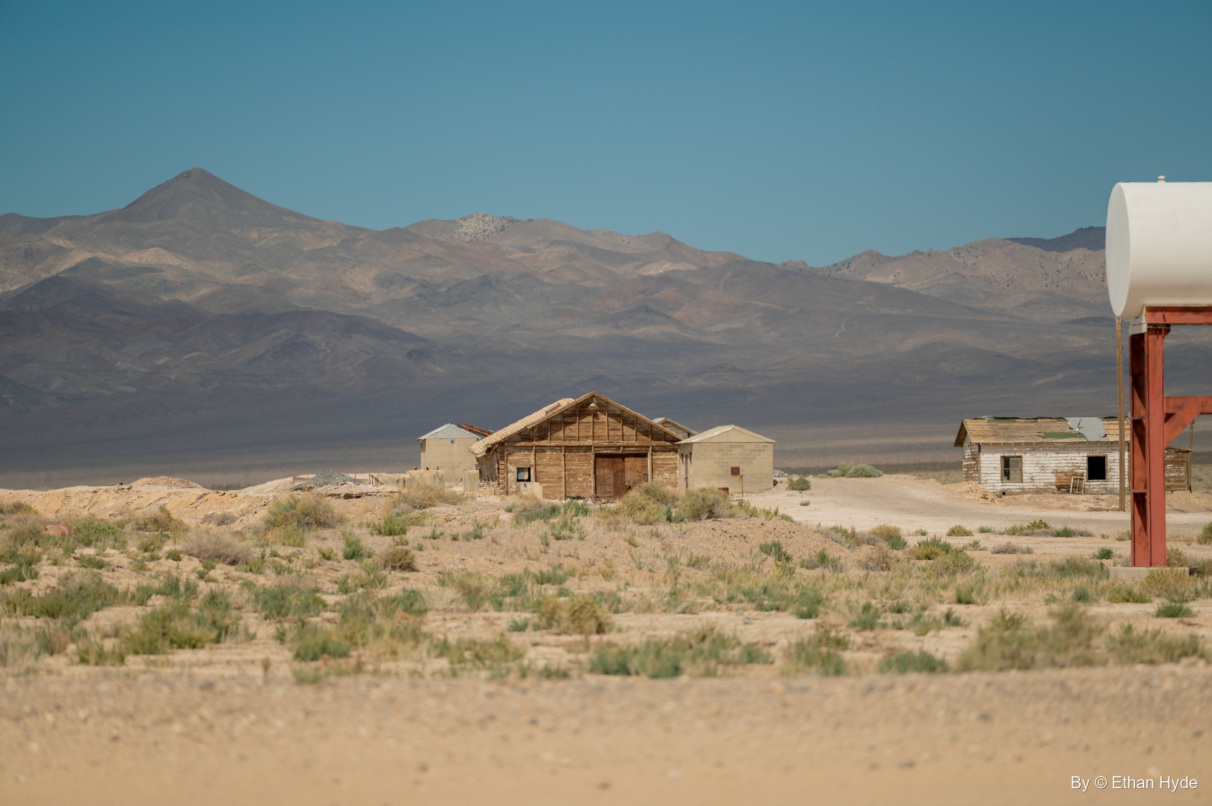 Weathered wooden houses sit in desert scrub below bare mountains, a red water-tank stand at the right edge.