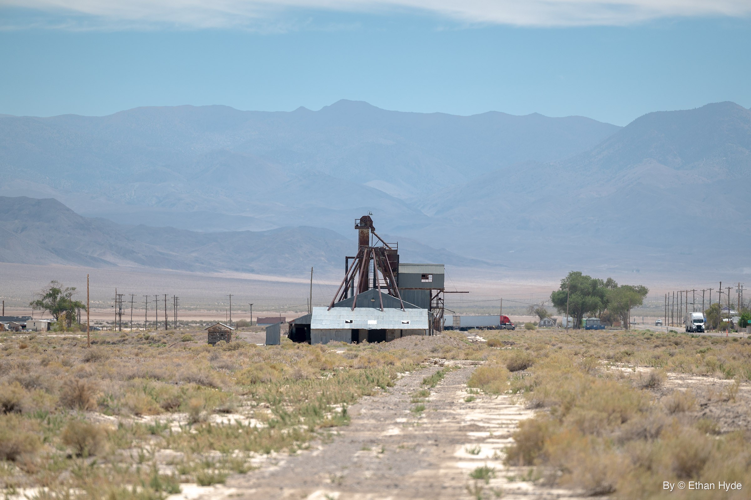 A weathered steel tower and a metal-roofed building stand in a sagebrush flat at the end of a dirt track, with trucks on a highway and hazy mountains behind.