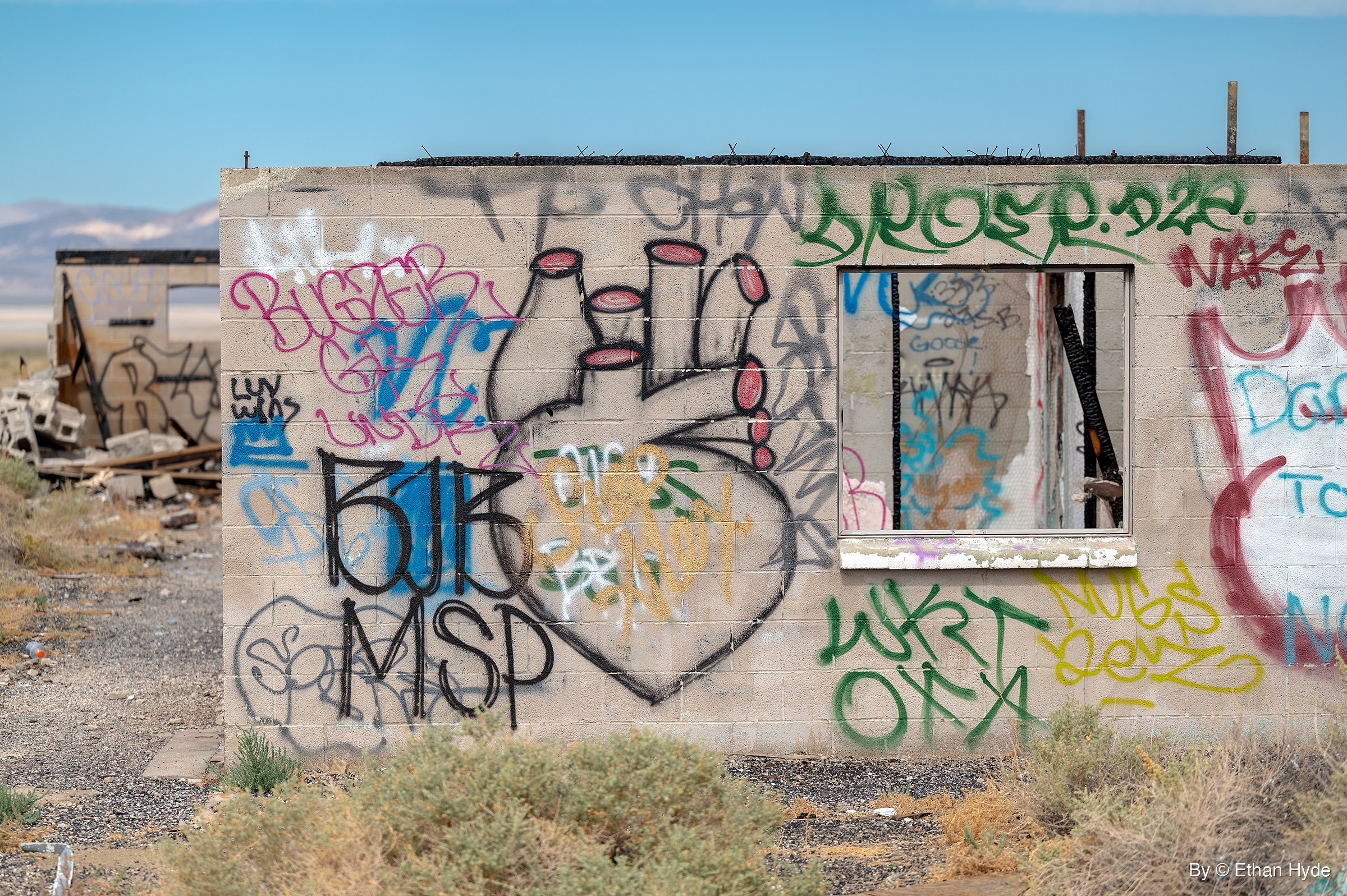 A cinderblock wall painted with a large anatomical heart outline and layers of tags, beside an empty window frame, with desert scrub in front.