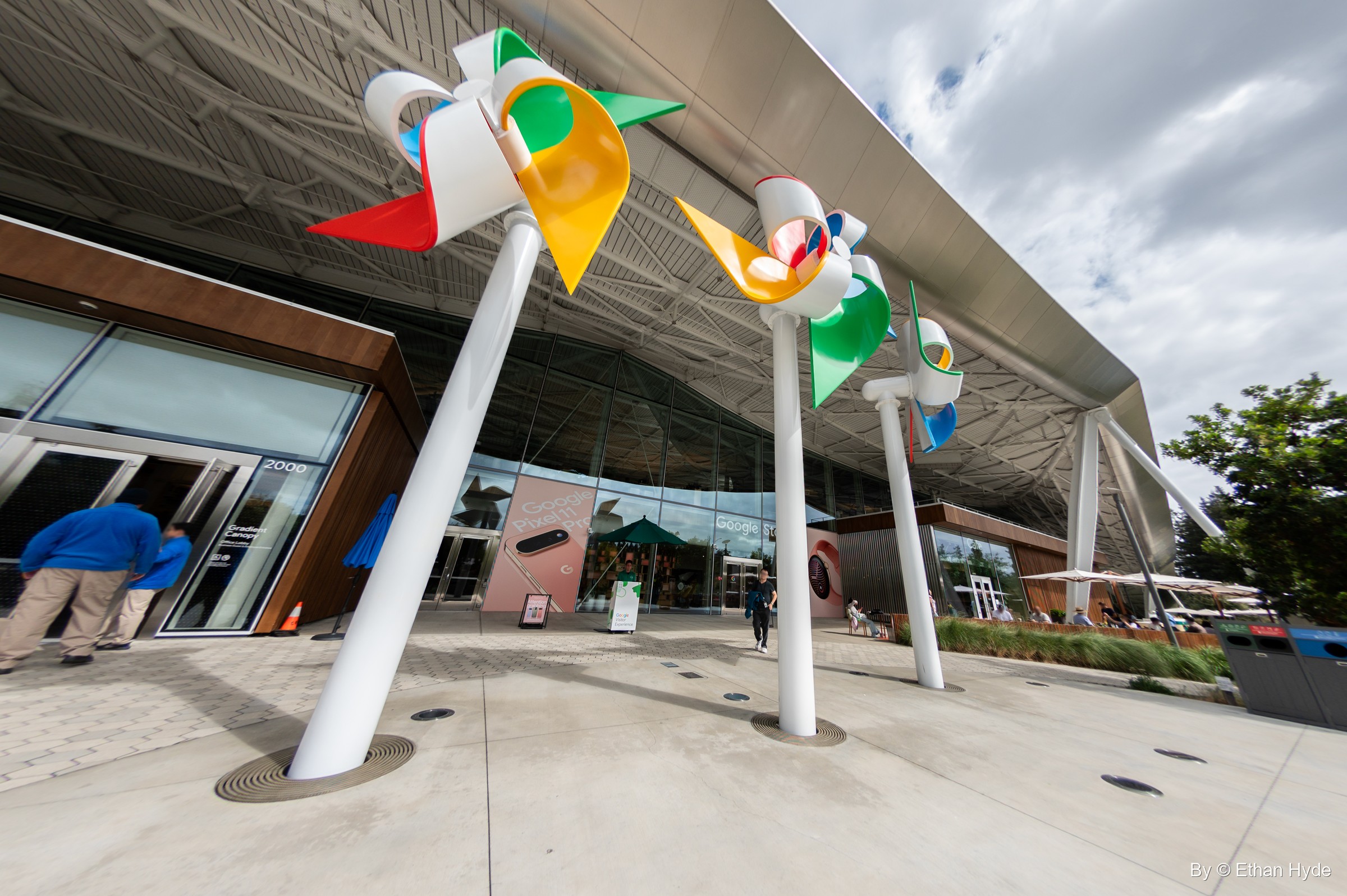 Giant pinwheels in Google colors on tall white poles stand at the entrance under Gradient Canopy's sweeping roof.
