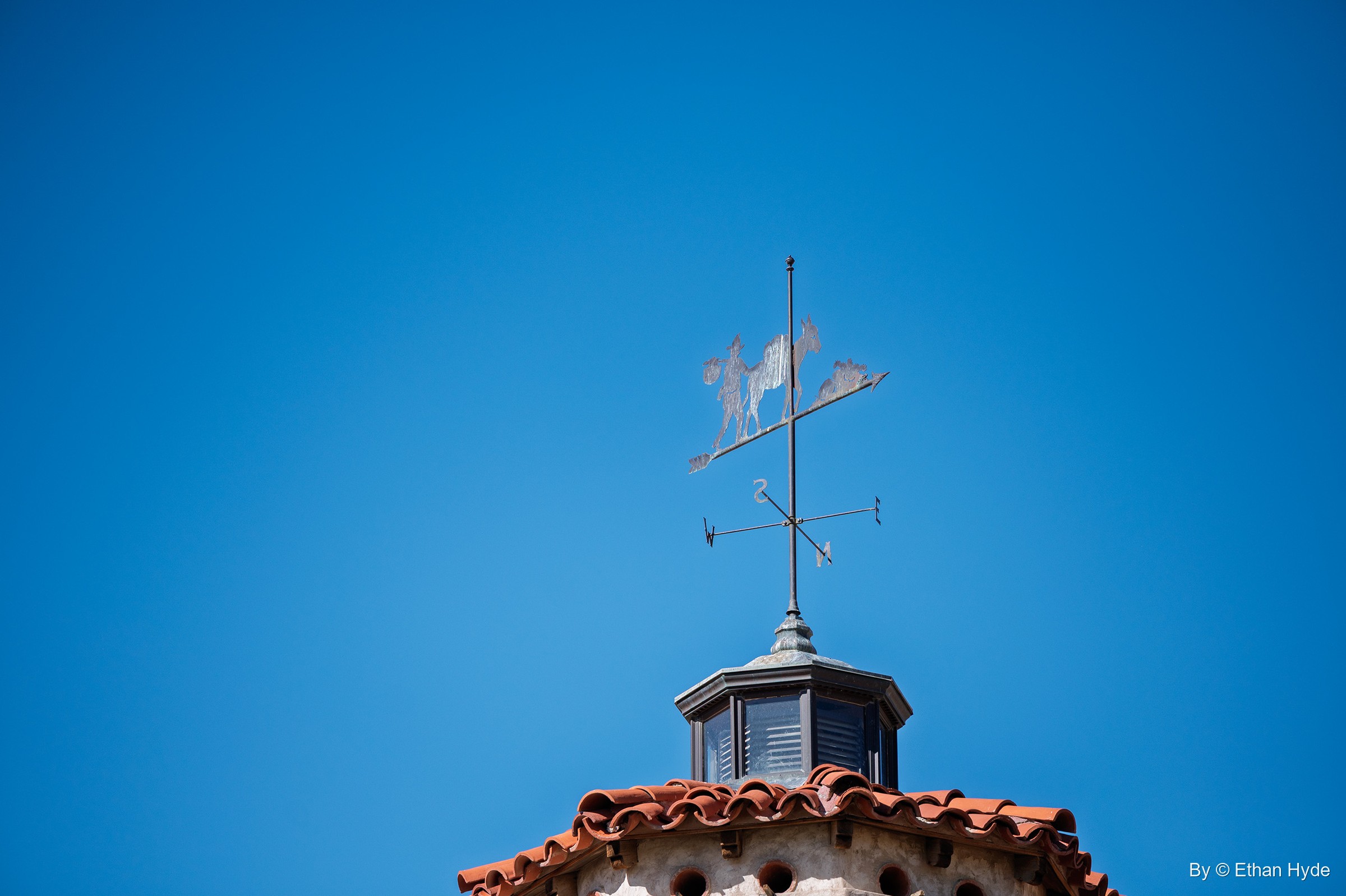 A weathervane showing a prospector carrying a bundle and leading a pack burro, atop a cupola at Scotty's Castle against a blue sky.